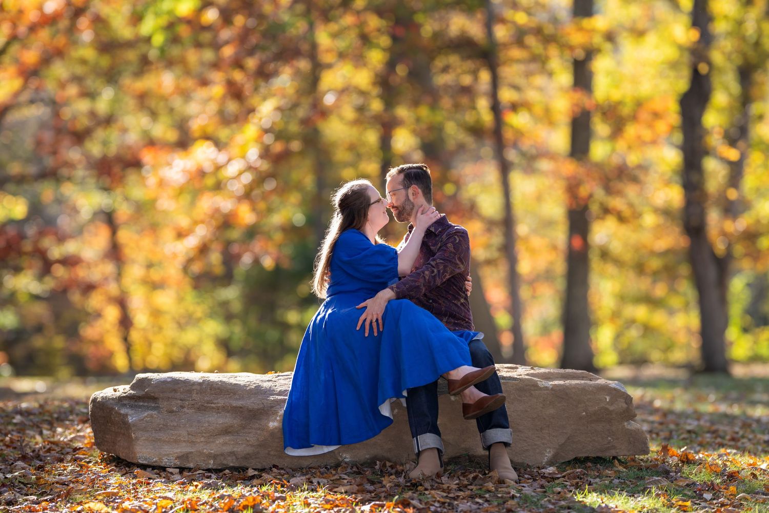 A couple sits together on a large rock surrounded by vibrant autumn foliage in a park setting.