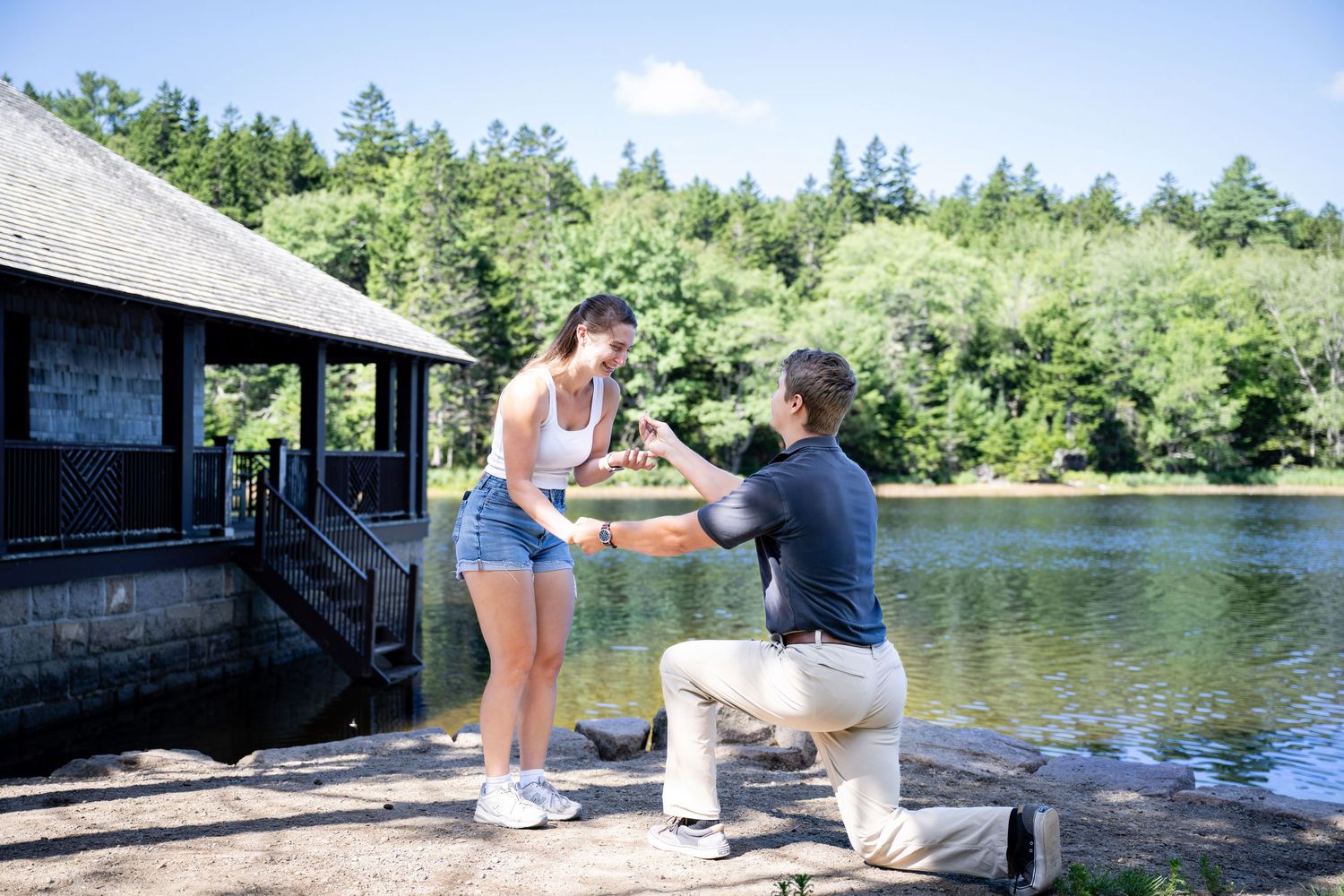 A romantic proposal scene unfolds on a wooden dock overlooking a serene lake surrounded by evergreen trees.