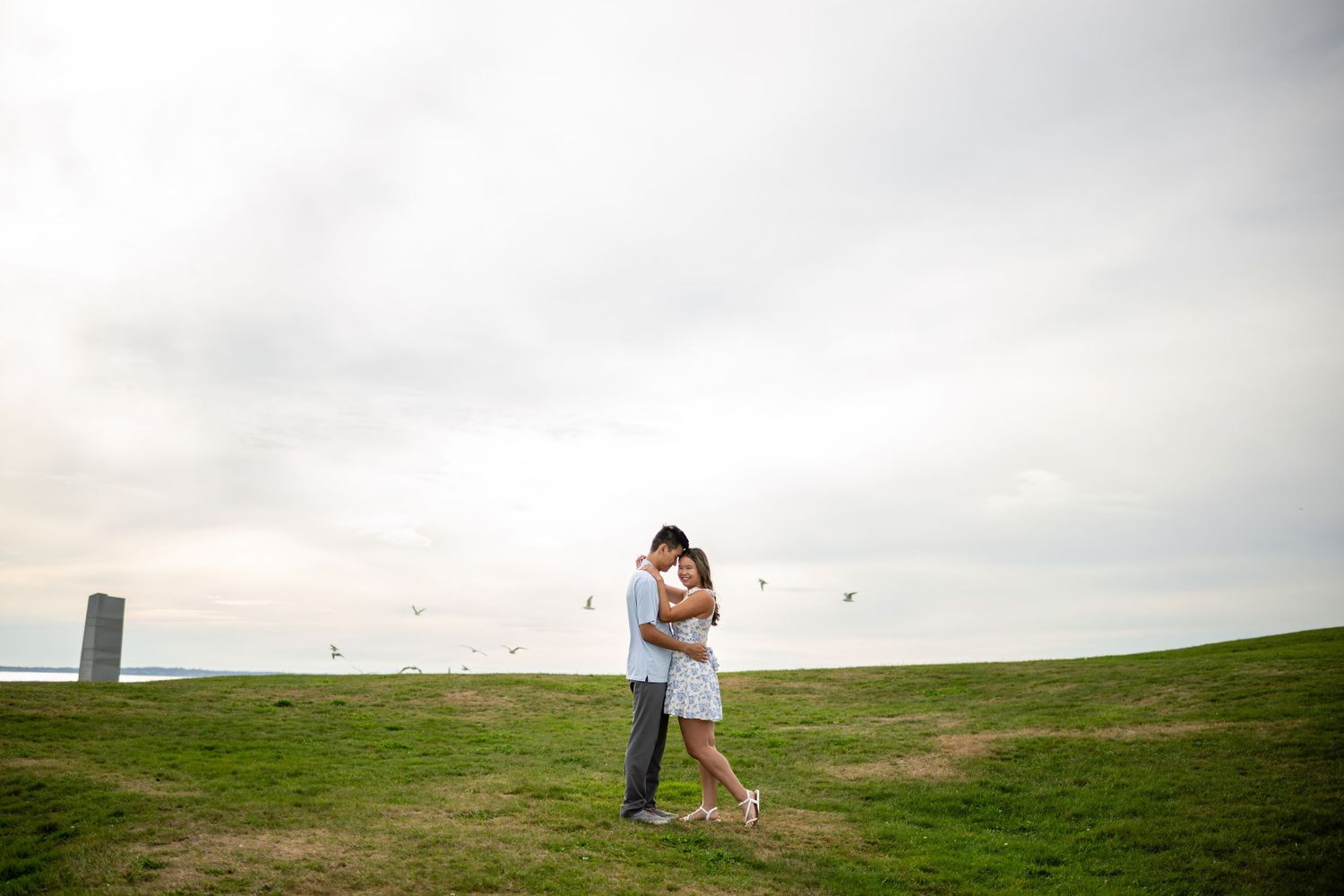 Engaged couple embracing at Brenton Point State Park in Newport, RI, with the rocky Atlantic coast in the background.