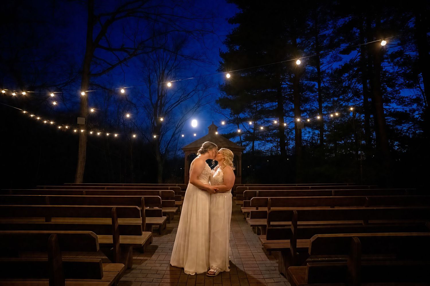 Same-sex couple shares a romantic kiss under the rustic pavilion at Crystal Lake in Middletown, with calm water and trees in the background.