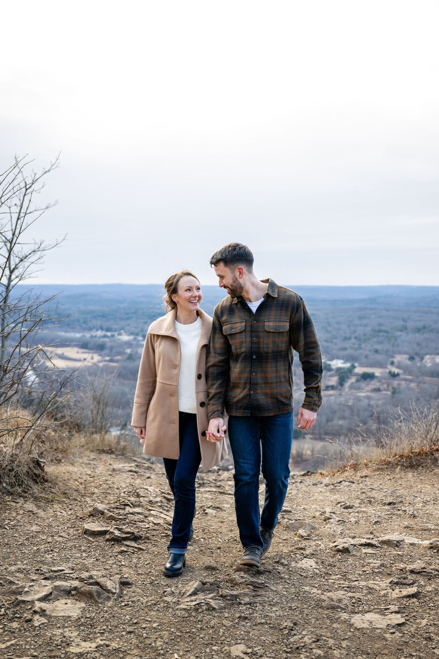A couple walks together on a hilltop trail overlooking a scenic mountain landscape on a cloudy day.