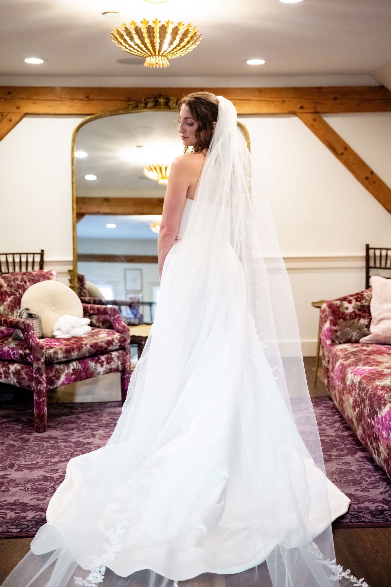 A bride in a flowing white wedding gown and veil stands in an elegant room with burgundy furniture and wooden beams.