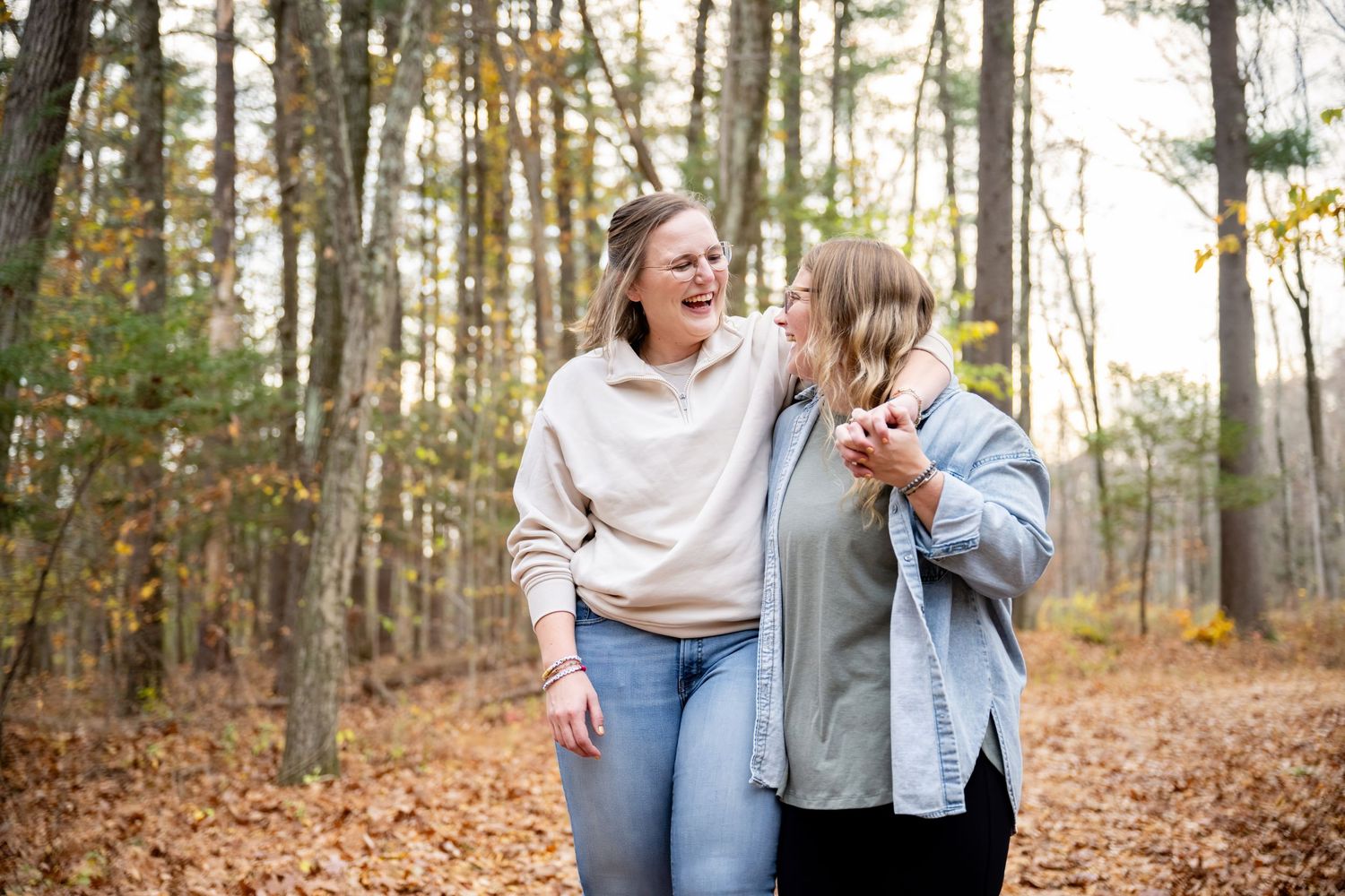 Two people laugh together while walking through a forest filled with autumn leaves and tall trees.