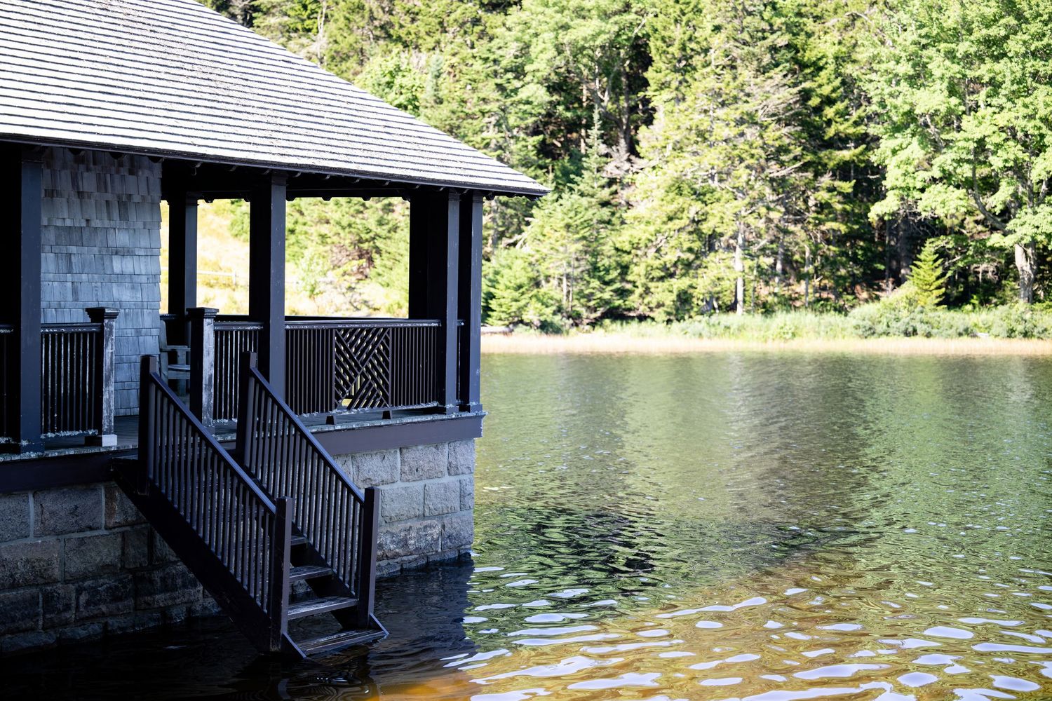 A black wooden boathouse with stairs sits on a tranquil lake surrounded by lush green trees on a sunny day.