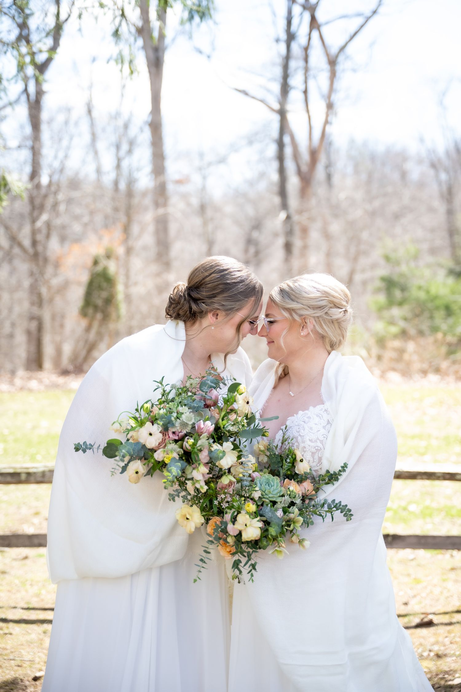 Two brides in white gowns and wraps touch foreheads, sharing a tender moment while holding a lush floral bouquet.