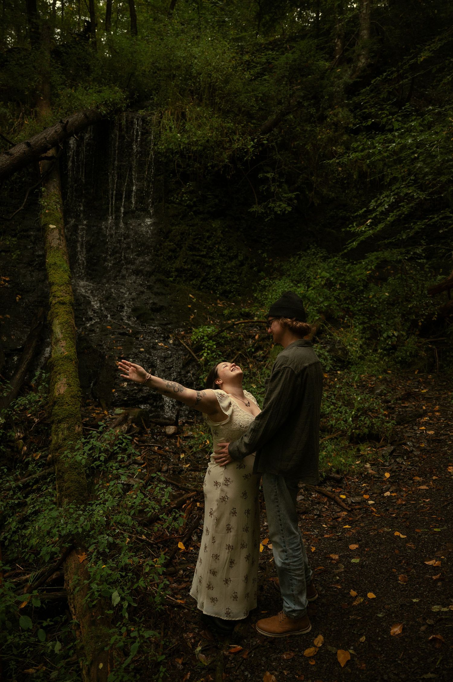 Couple embraces in lush forest near waterfall, woman in floral dress reaches up while man in hat holds her close.