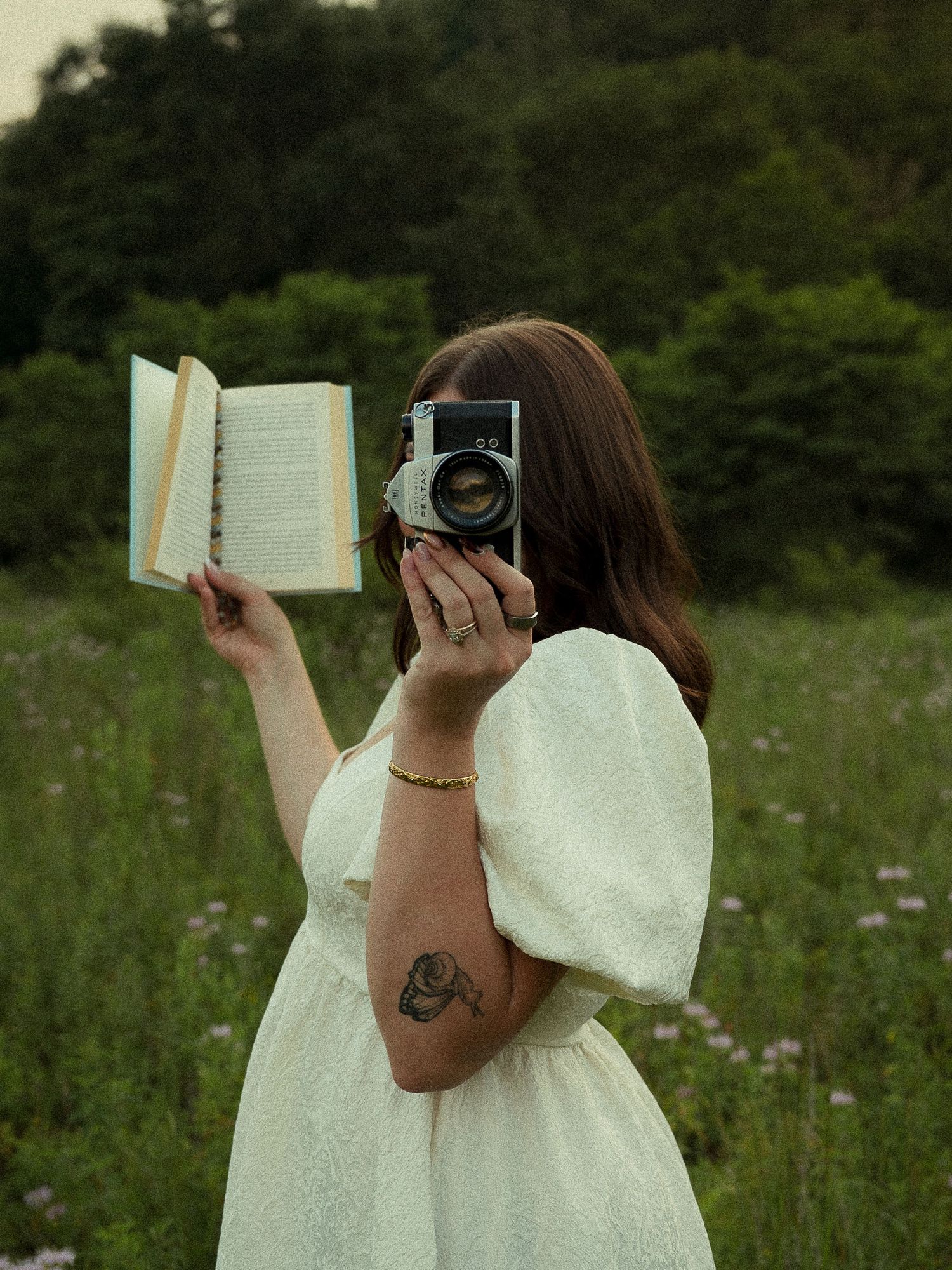 Woman in white dress holds book and vintage camera in a wildflower field with lush green trees behind her.