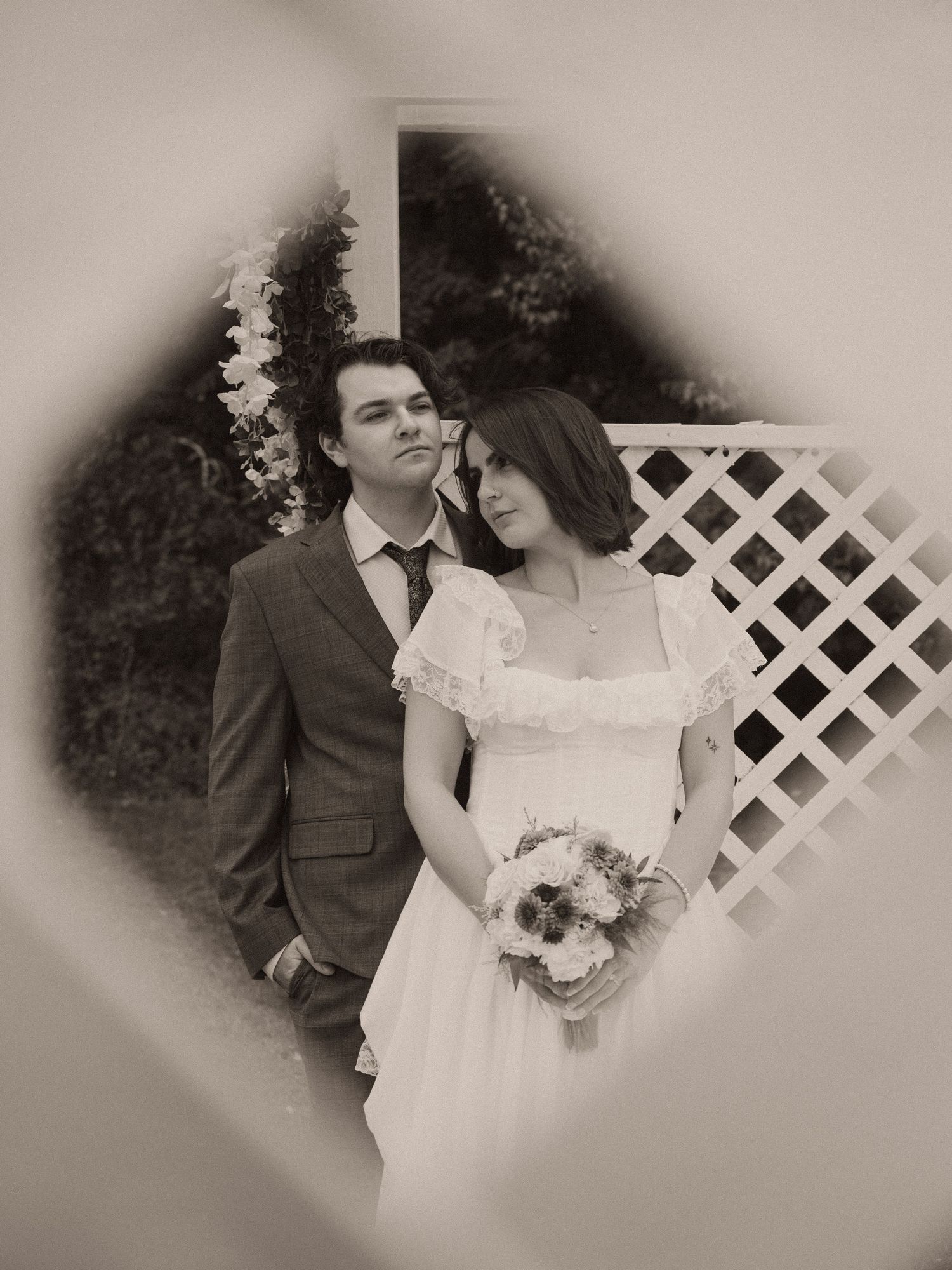 Sepia-toned wedding portrait of bride holding bouquet leaning on groom in suit, framed by heart-shaped vignette.
