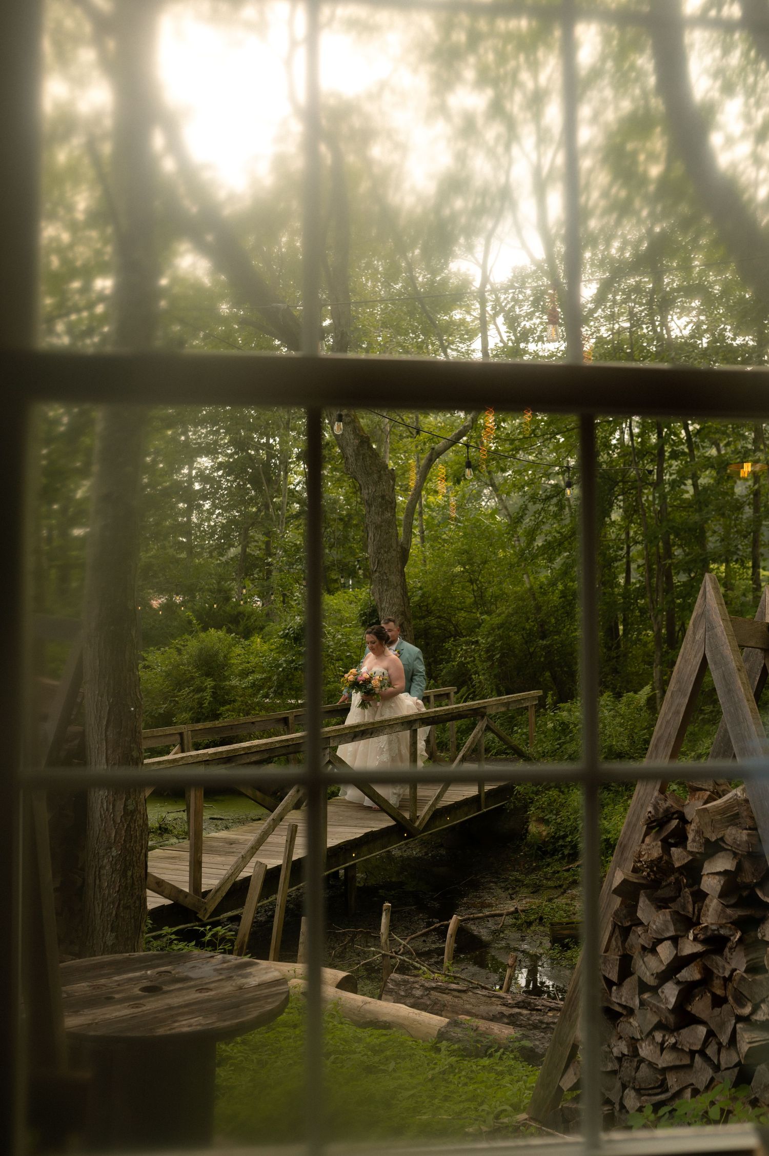 A rustic wooden bridge in a forest setting with stacked firewood along the sides.