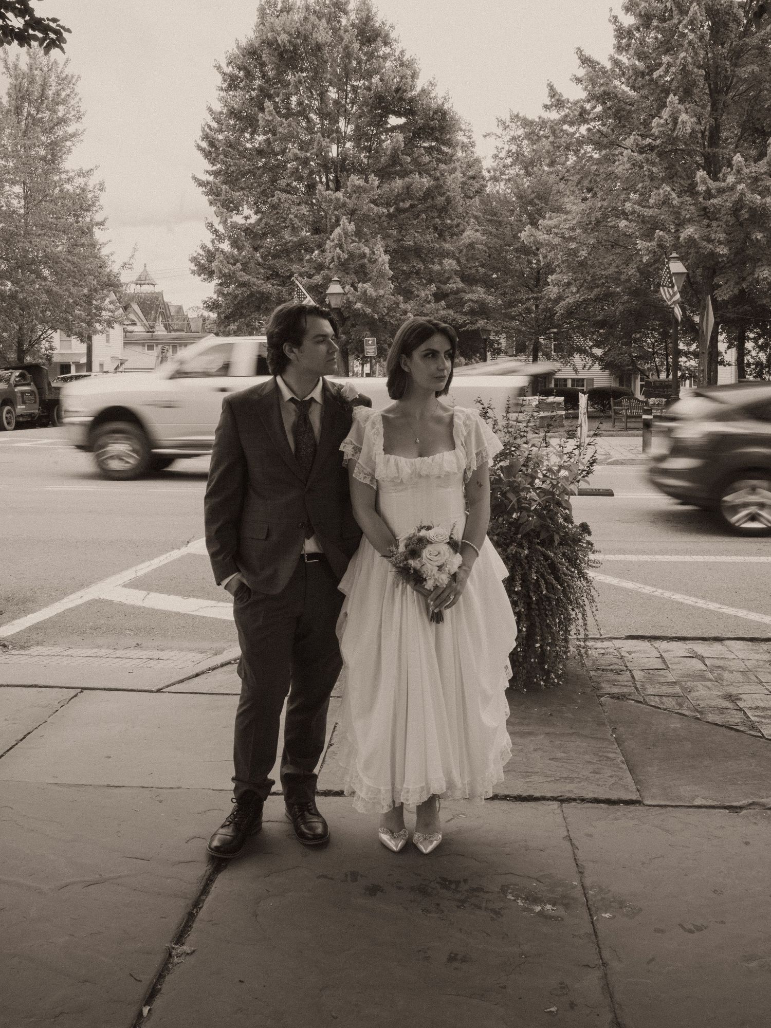 Black and white photo of a bride and groom standing on a sidewalk with trees and passing cars in the background.