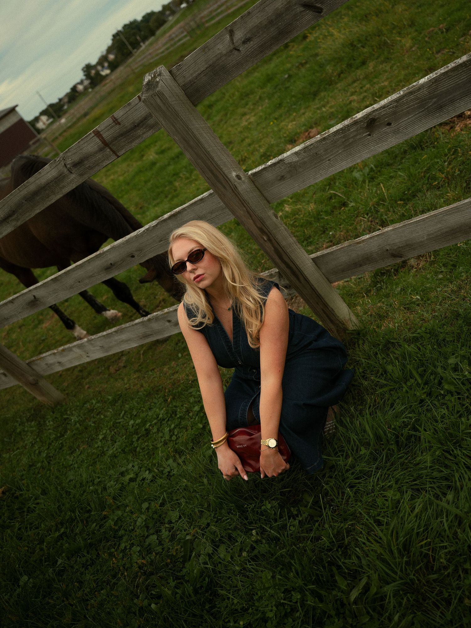 Blonde woman in sunglasses crouching on green grass by wooden fence with horses grazing in background.