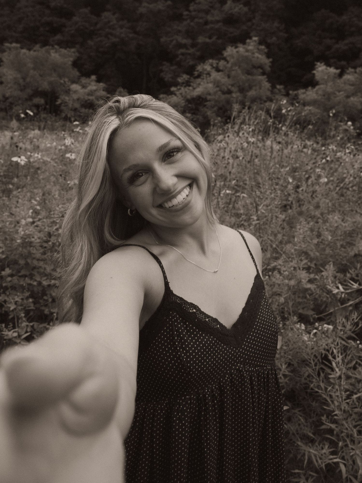 Smiling blonde woman in a floral field reaching toward camera in a black and white selfie-style photo outdoors.