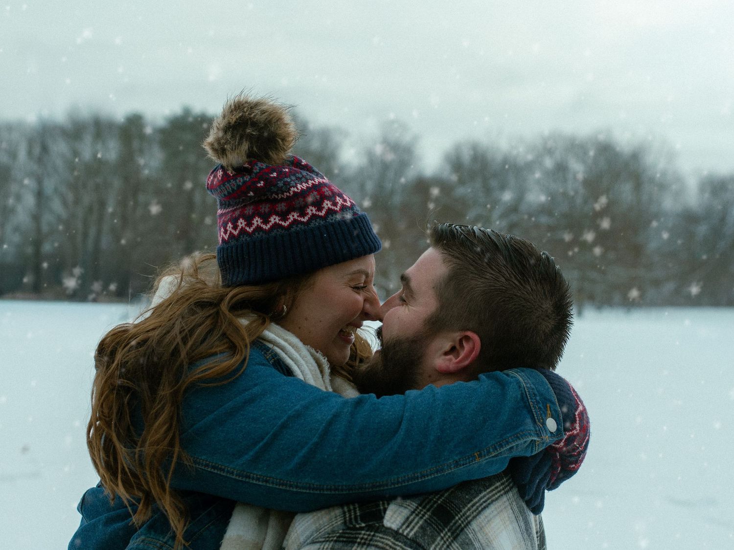 A couple embraces and laughs together in a snowy winter scene while one wears a knit pom-pom hat.