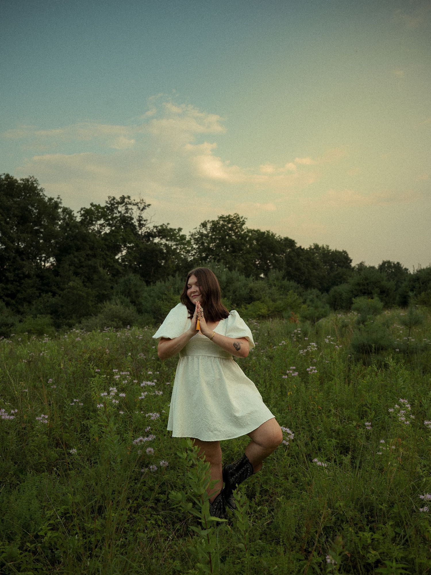 Woman in white dress and boots standing in a wildflower meadow with hands pressed together, trees and cloudy sky behind.