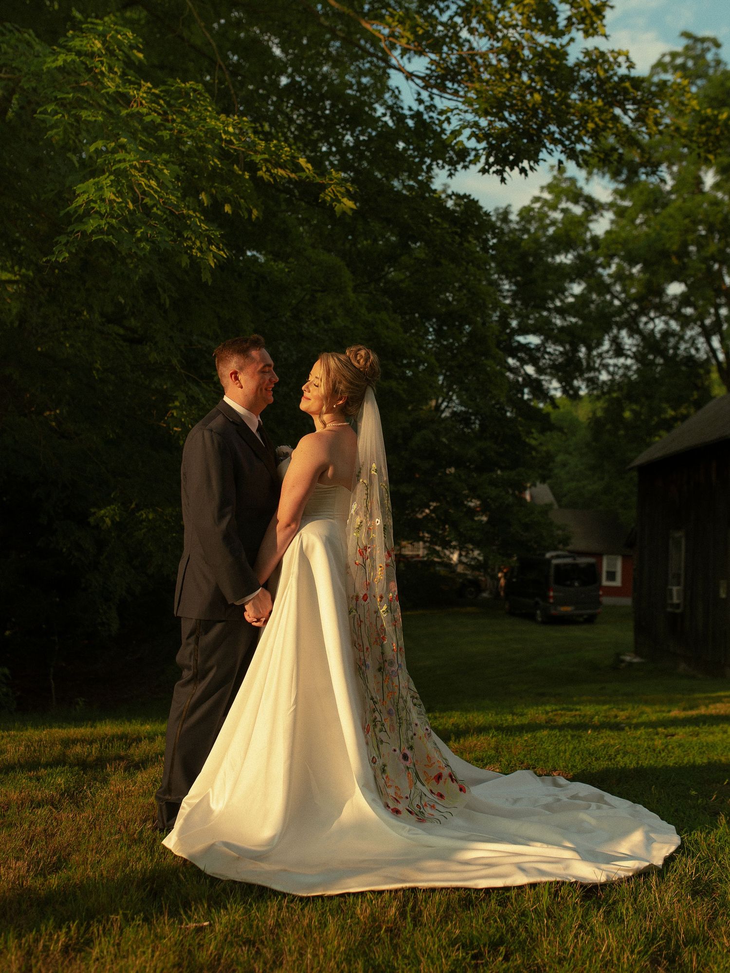 Bride and groom holding hands in golden sunset light, standing on green grass with lush trees in background.