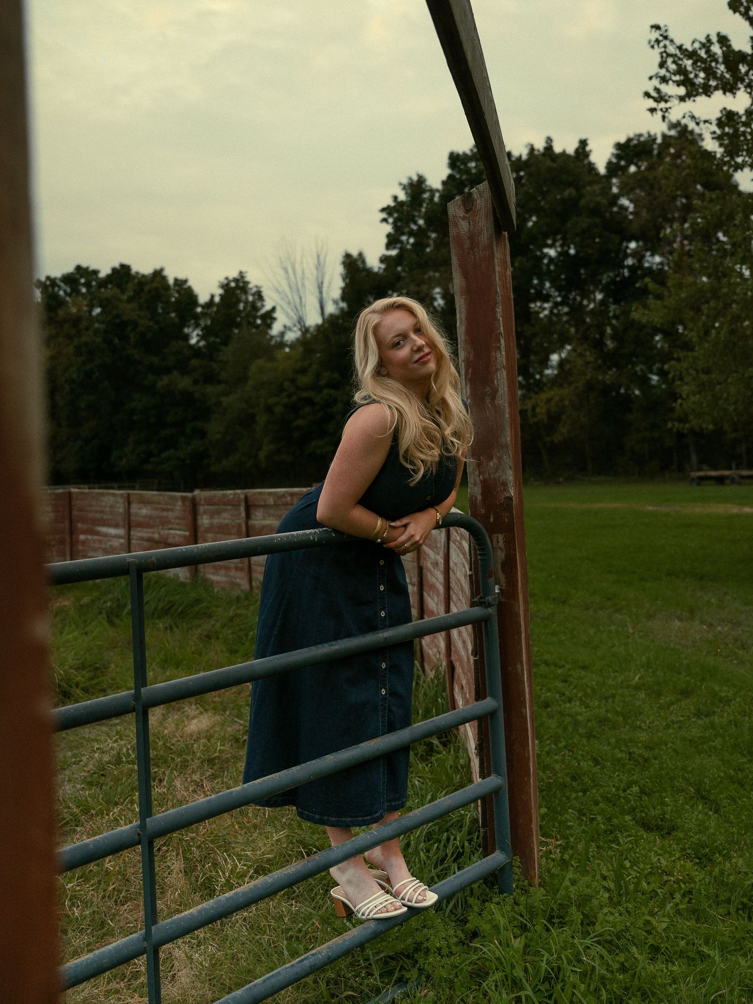 Blonde woman in dark teal midi dress leaning on metal farm gate in green field with trees.