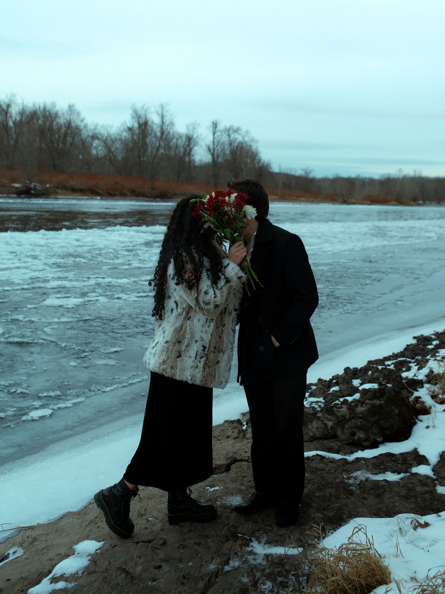 Two figures stand together on a snowy riverbank in winter with bare trees and frozen water in the background.
