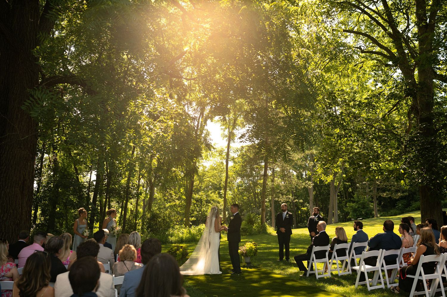 A sun-drenched wedding ceremony site with white chairs arranged in rows among trees.