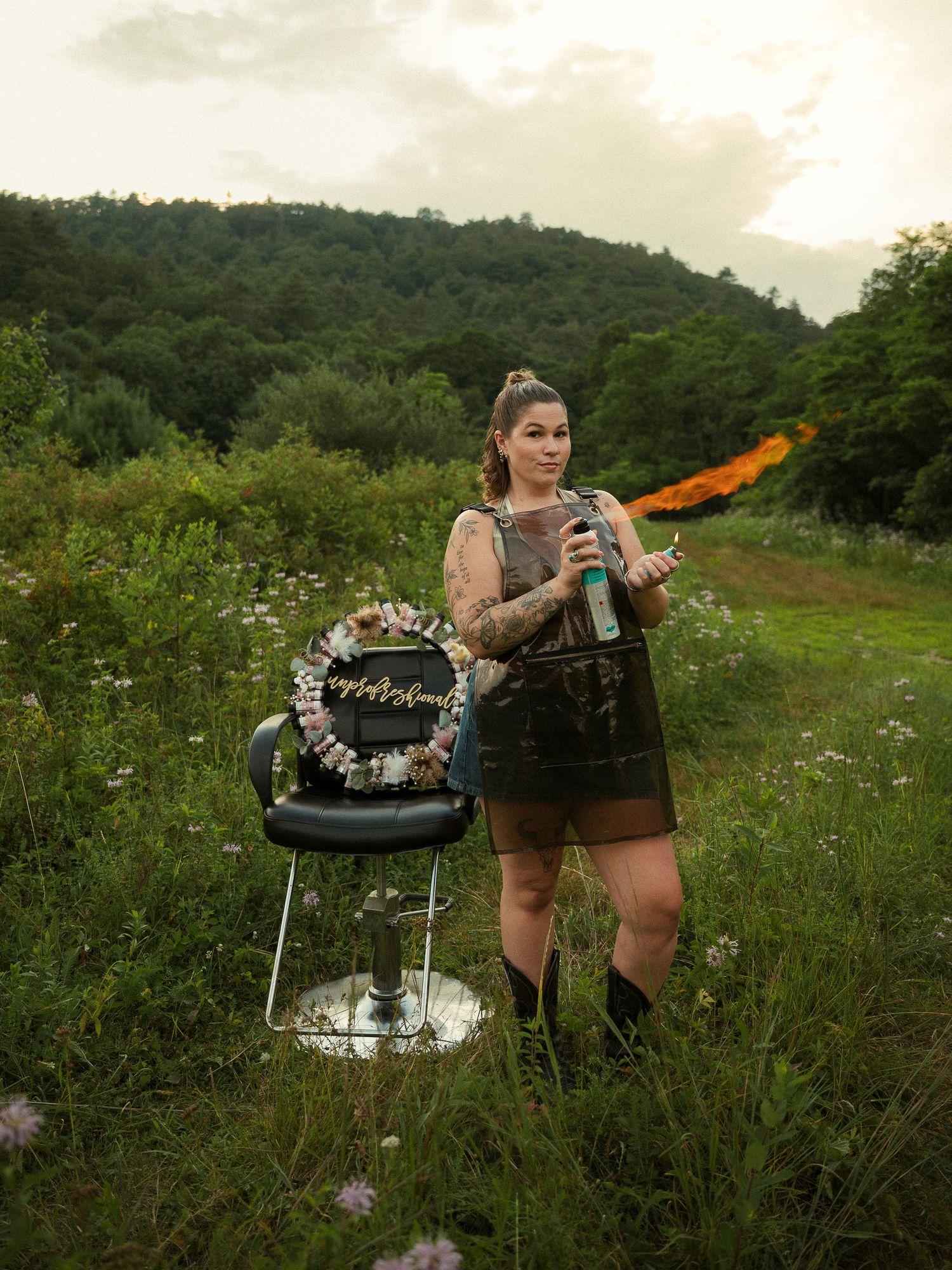Tattooed woman holding hair tools stands beside a salon chair in a lush green field with a smoke flare behind her.