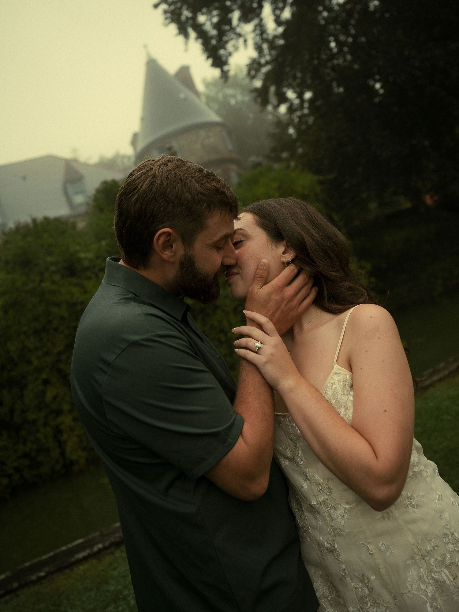 A couple shares a romantic almost-kiss outdoors, the woman in a lace dress and man in green shirt, with a misty castle backdrop.