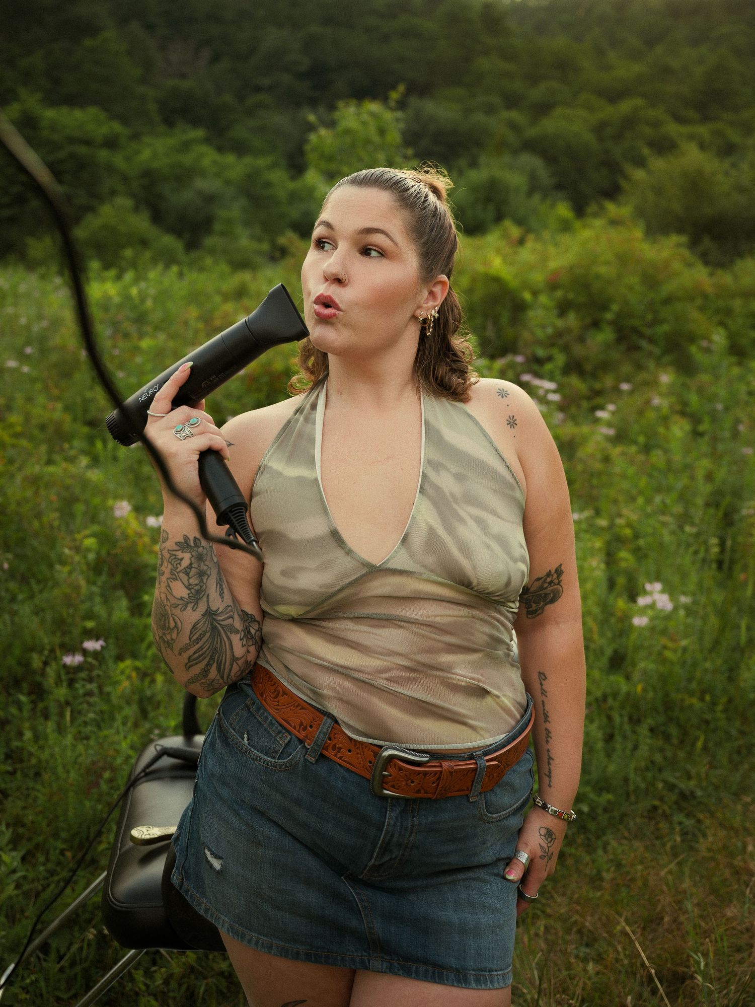 Woman with tattoos holding a megaphone in a lush green field, wearing a sheer top and denim skirt.