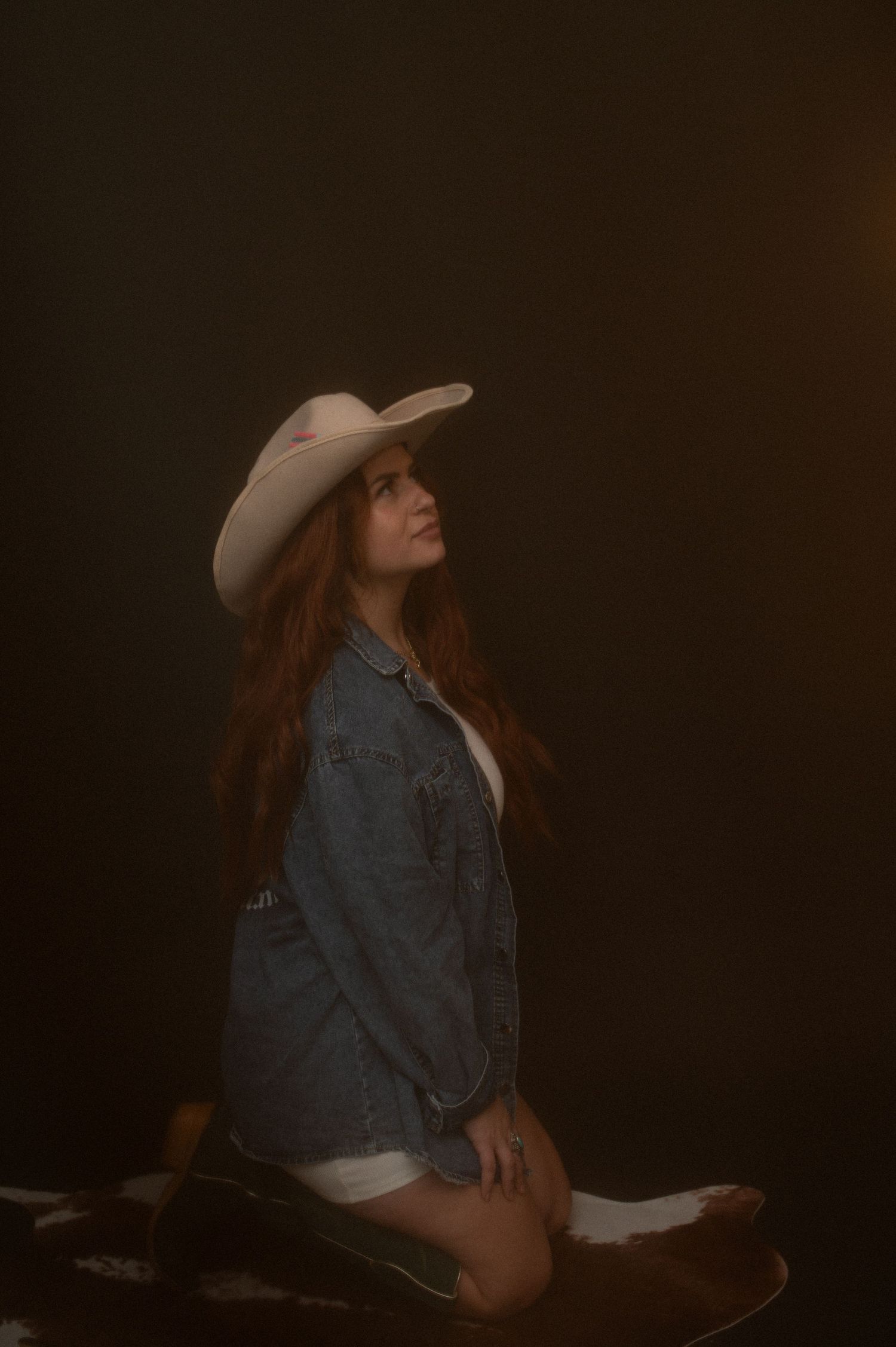 A person wearing a denim jacket and wide-brimmed hat poses in dramatic low-key lighting against a dark background.