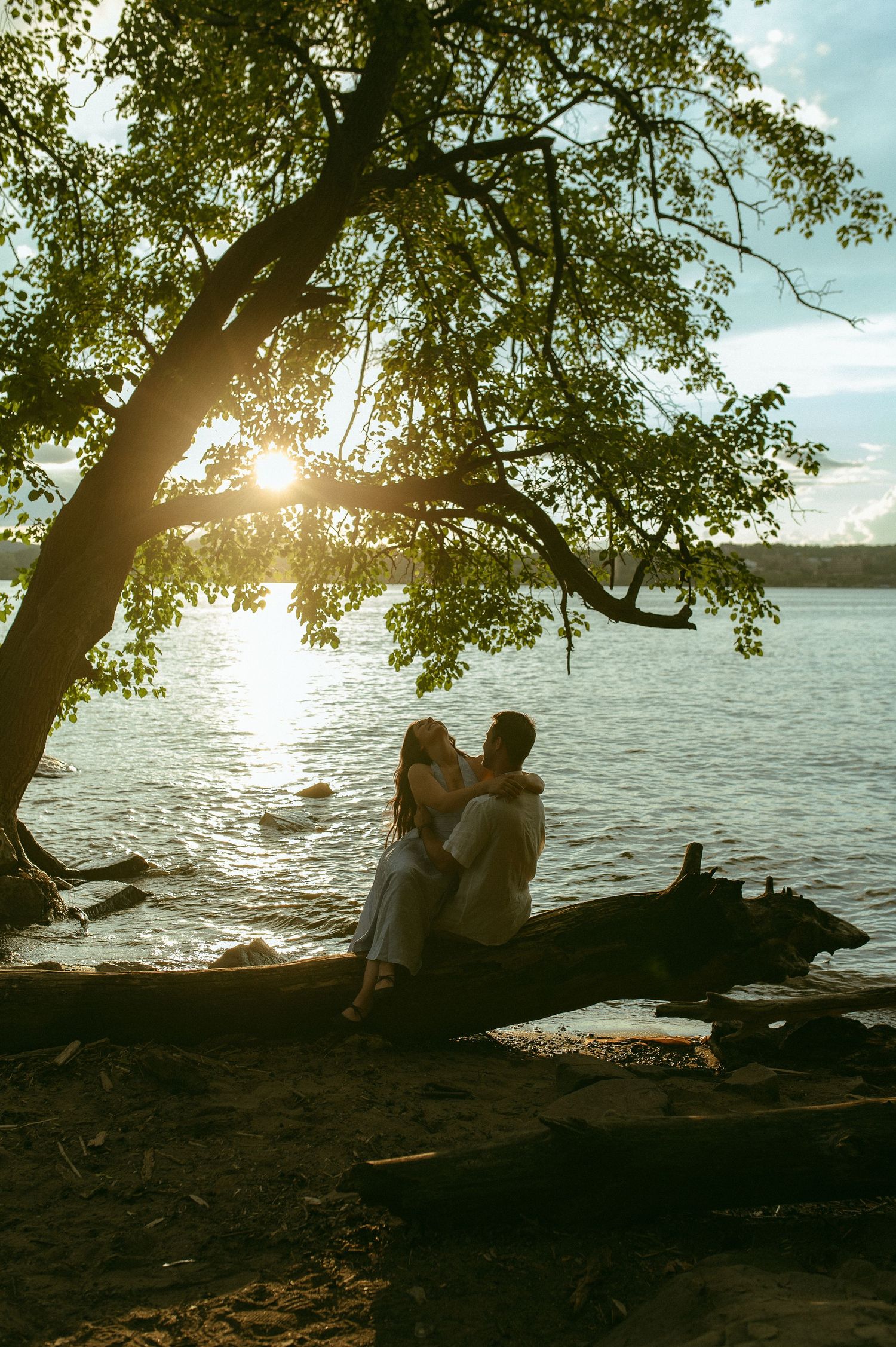 Couple sitting on a fallen log by a lake under a large tree at golden sunset.