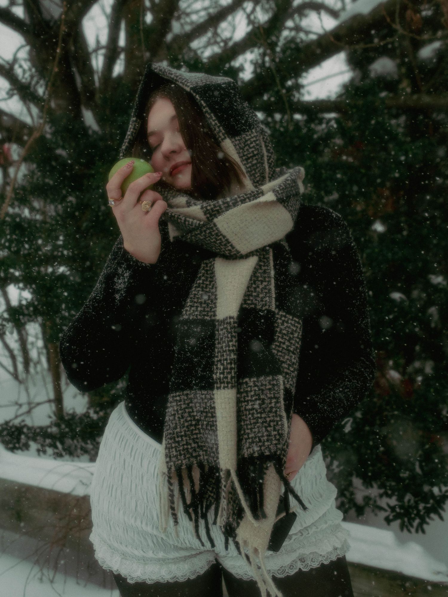 Person wearing a black and white patterned scarf and winter coat poses playfully in snowy winter scenery.