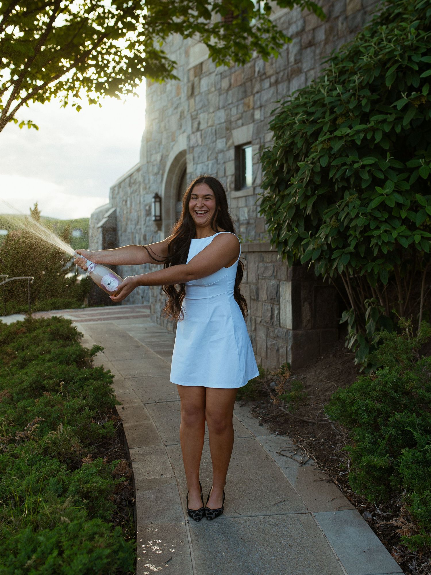 Young woman in white dress spraying champagne on a stone pathway outside an elegant building at sunset.