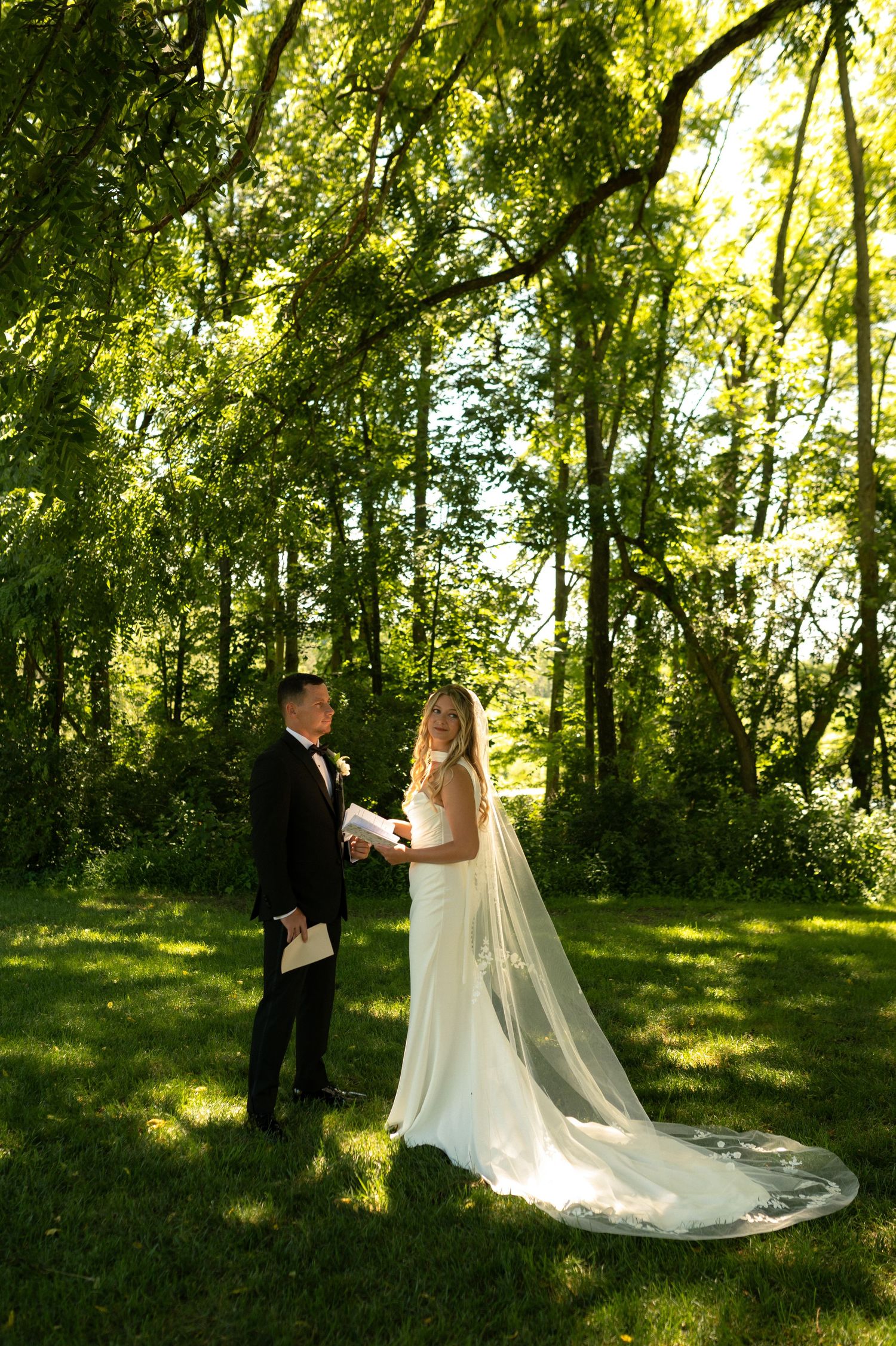 Wedding couple standing in a sunlit forest clearing with a flowing white dress and veil.