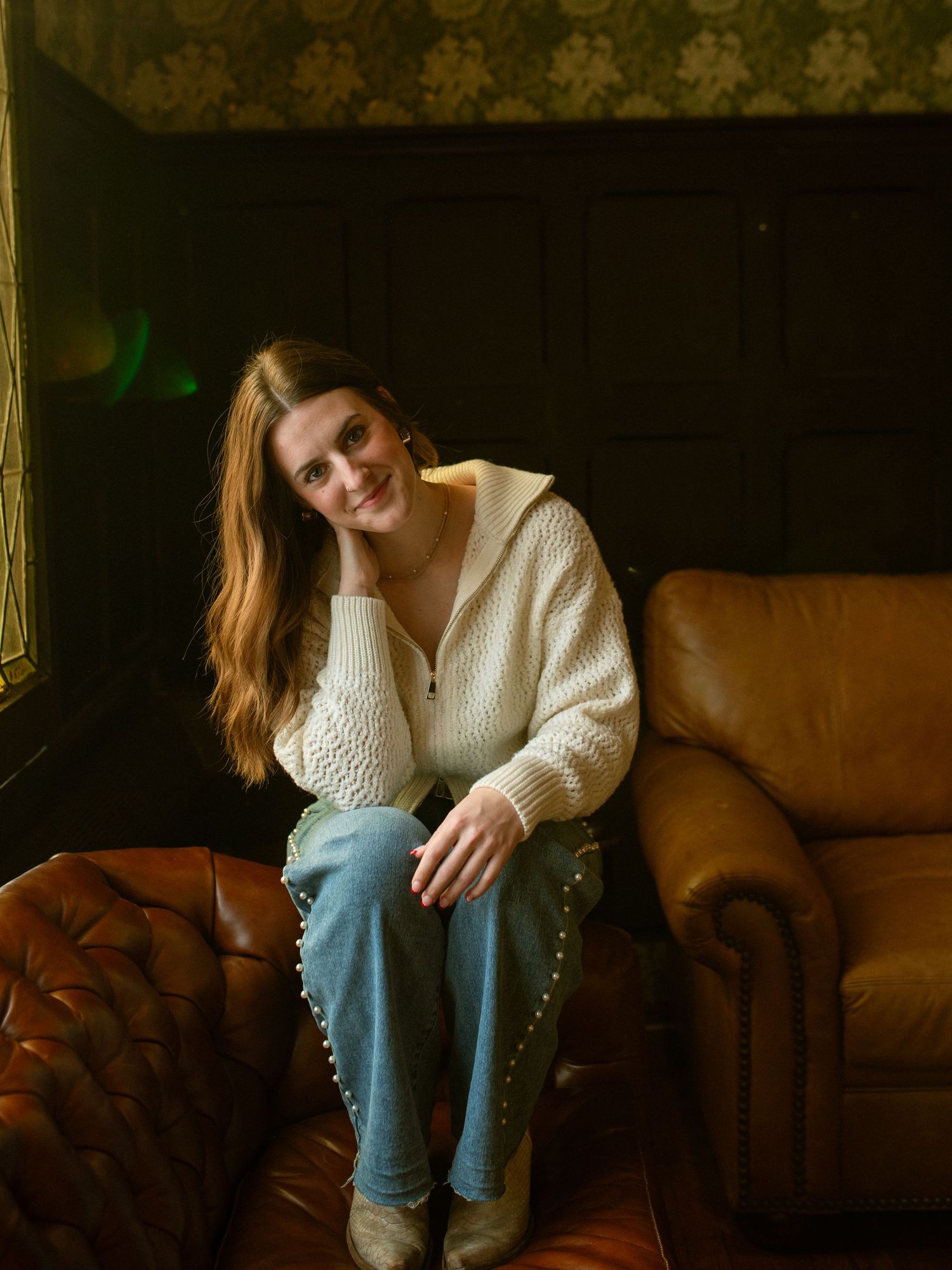 Person in white sweater and blue jeans sitting on brown leather couch in moody lighting.