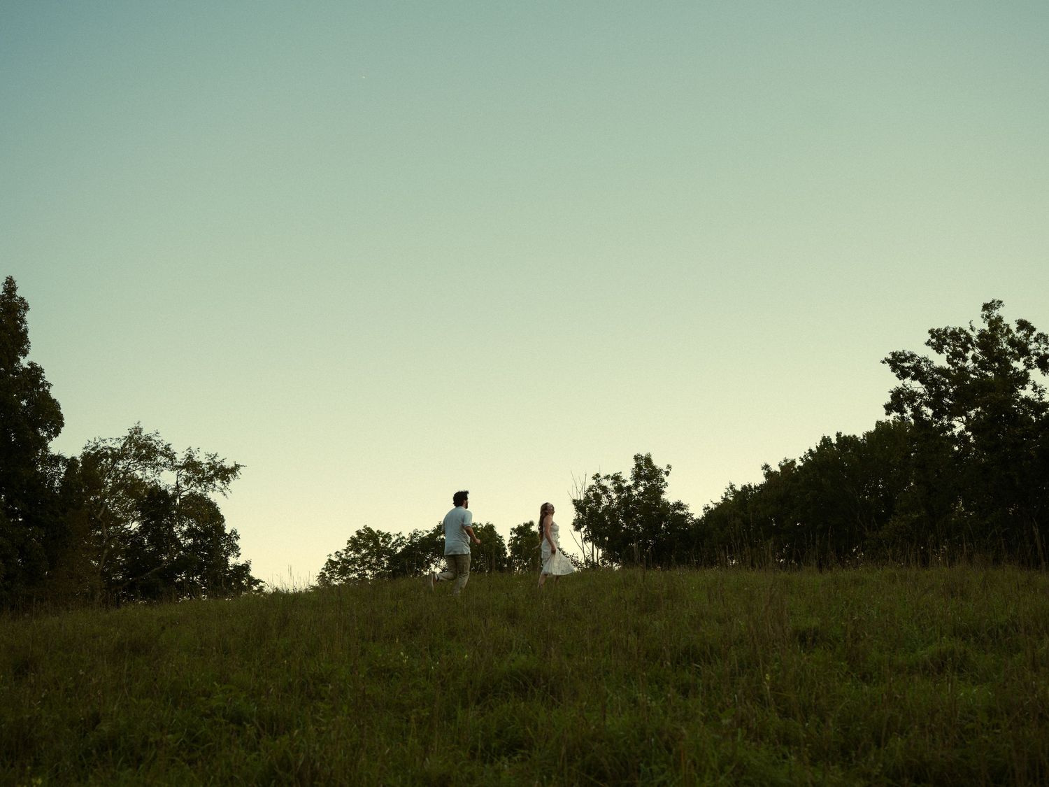 Couple in wedding attire walking hand in hand across a green hillside at dusk, surrounded by trees.