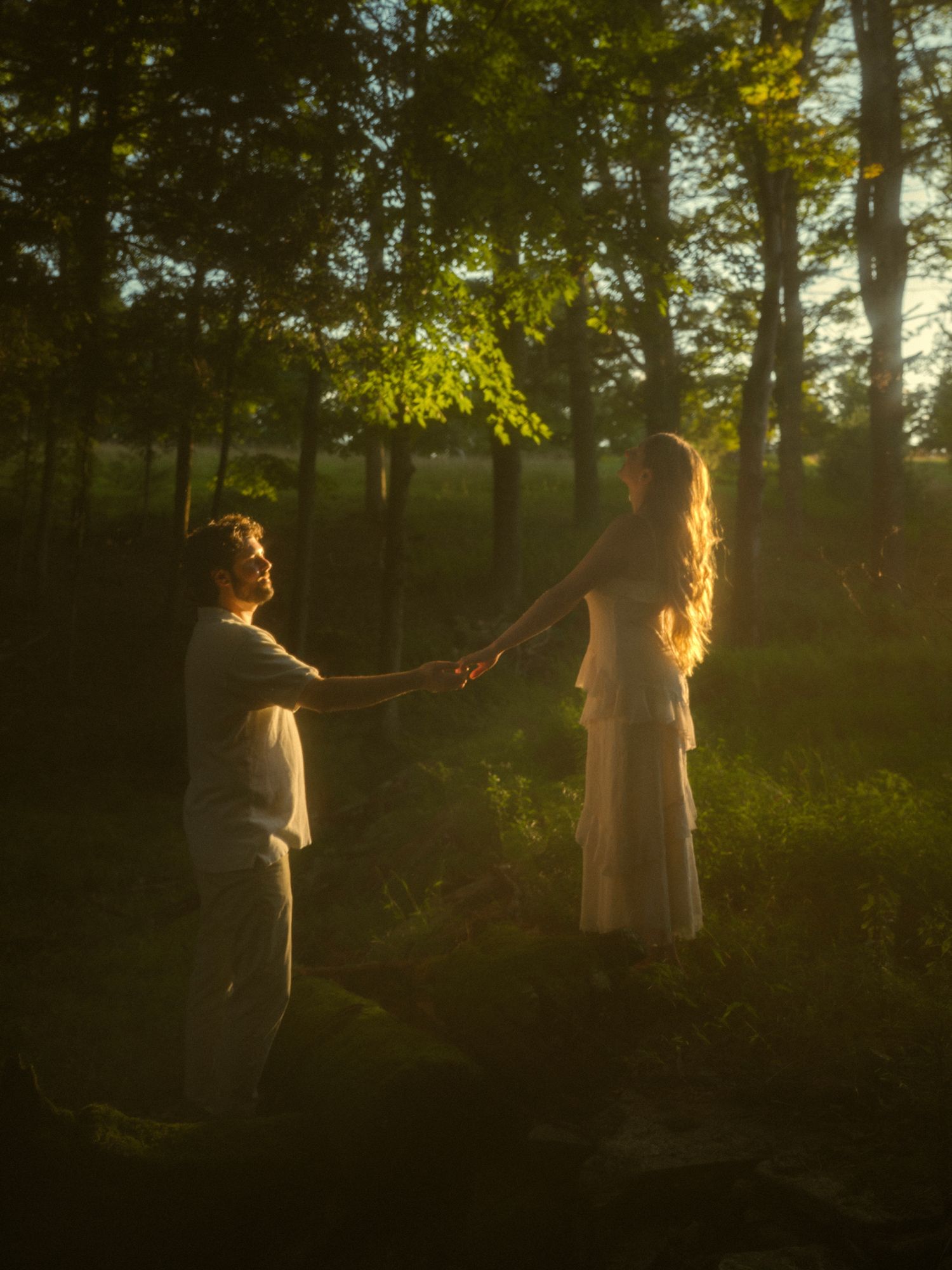 A couple holds hands in a sunlit forest clearing at golden hour, woman in white dress with long blonde hair.
