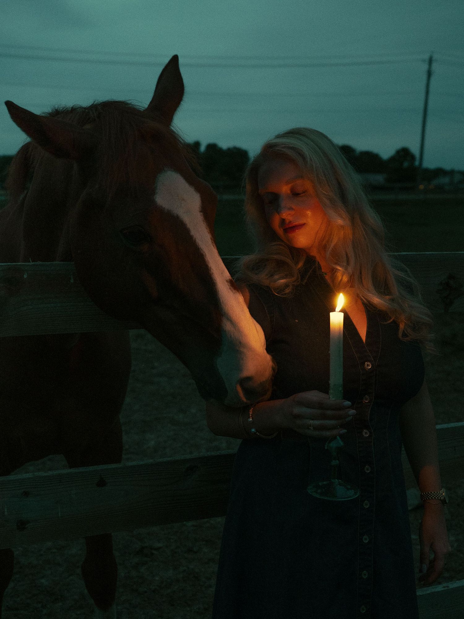 Woman with long blonde hair holds a lit candle beside a brown horse at dusk near a wooden fence.