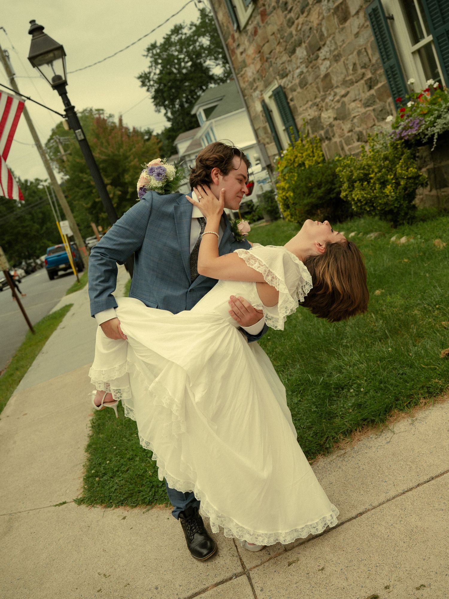 Groom dipping bride in white wedding dress on sidewalk, sharing romantic kiss outdoors near stone building.