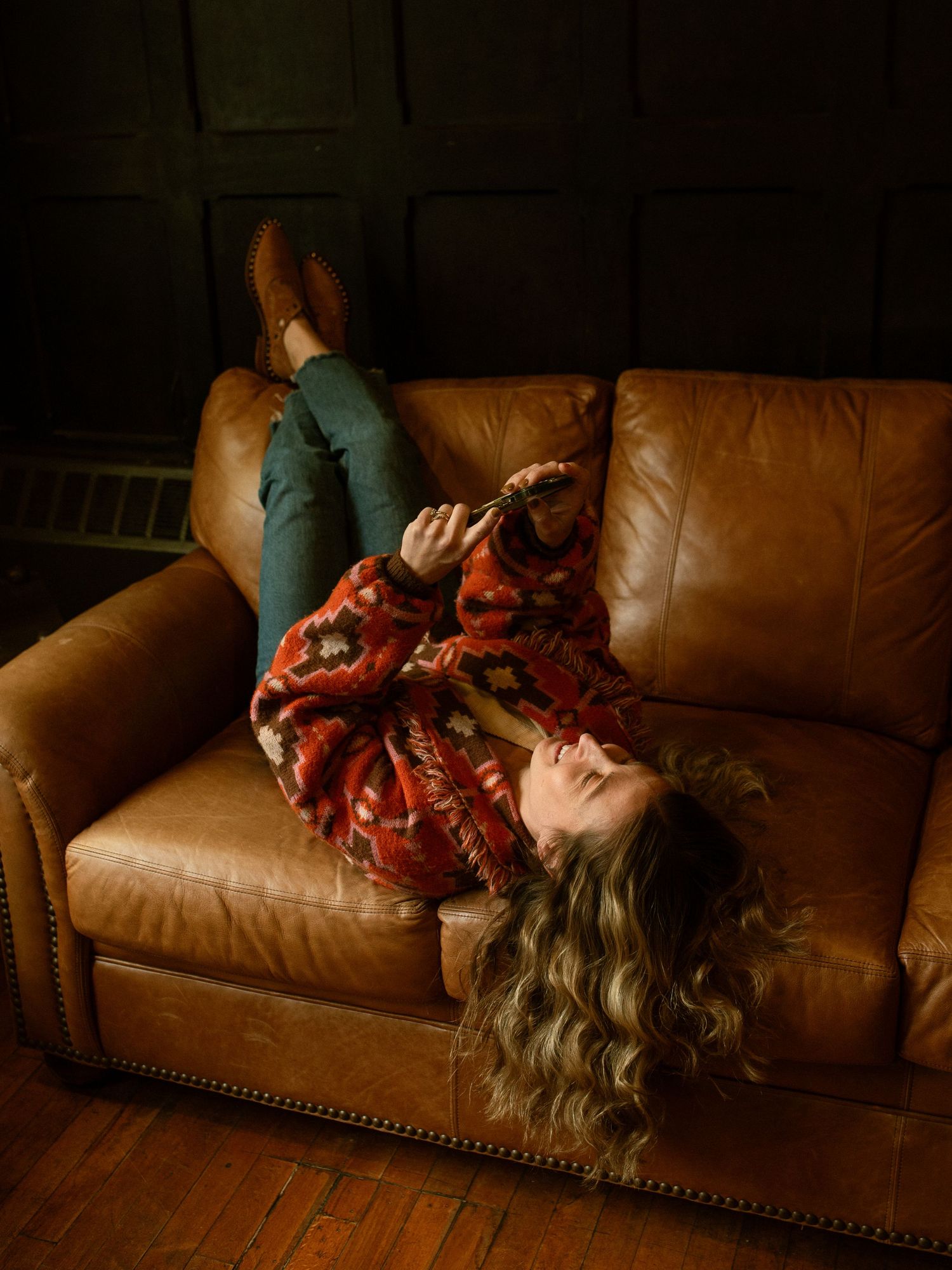 Person relaxing on brown leather couch wearing red patterned sweater and green pants while looking at phone in dark room.