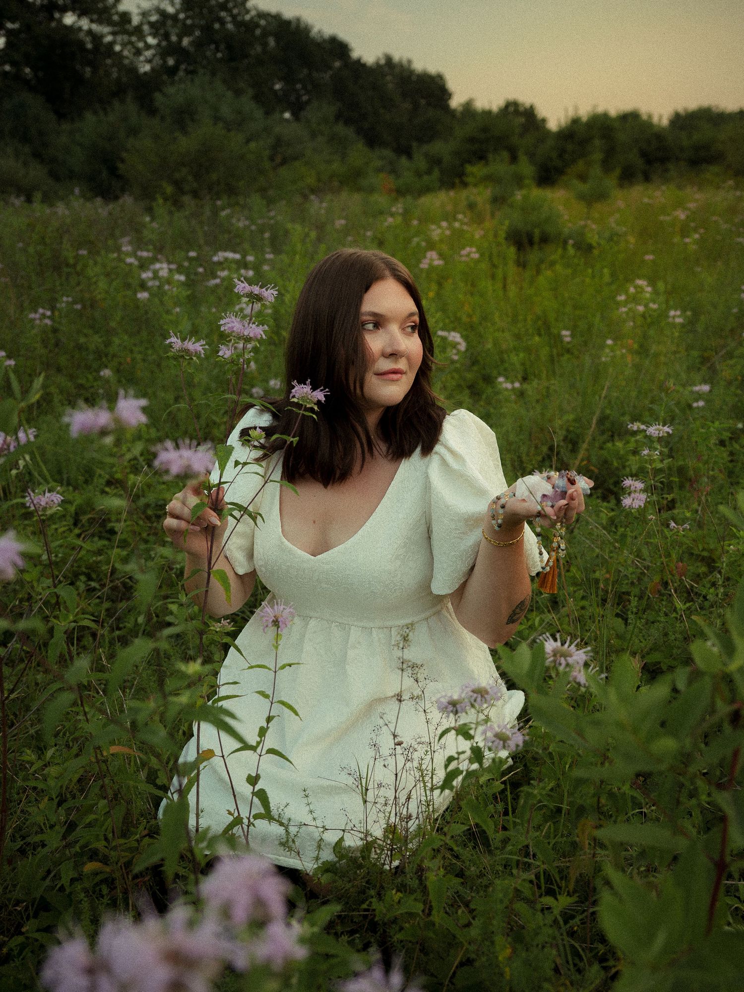 Young woman in white dress sitting in a wildflower meadow holding purple flowers at dusk.