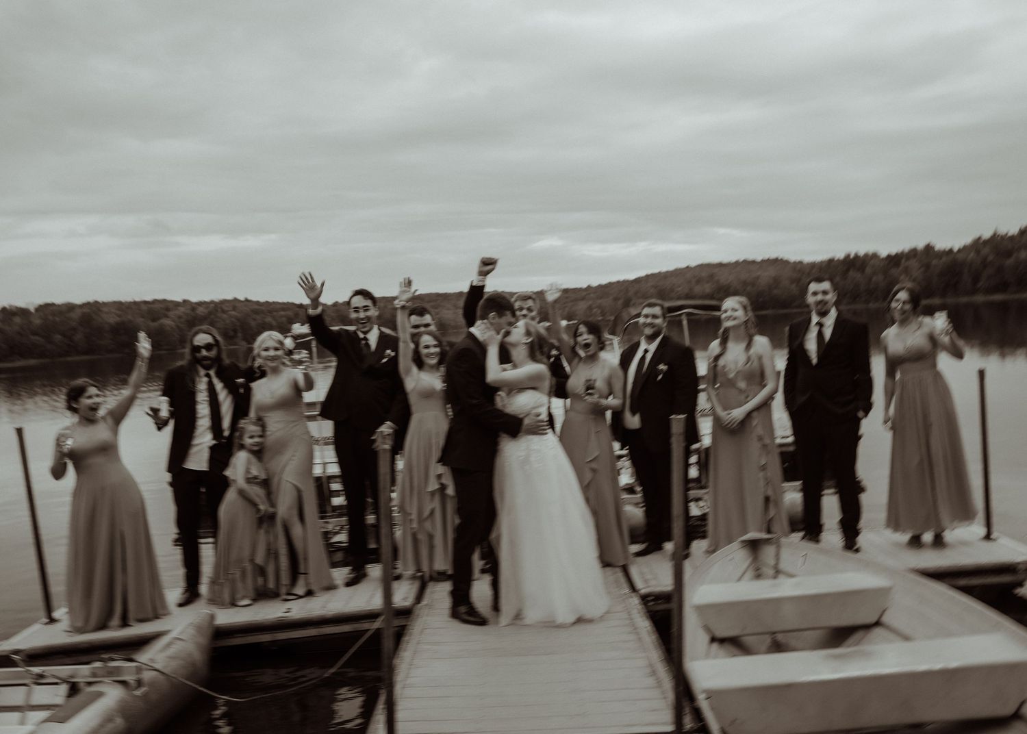 Wedding party gathers on a lakeside dock under cloudy skies.