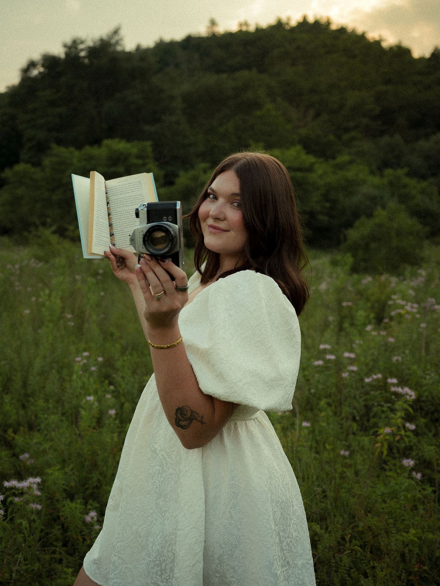 Young woman in white dress holding vintage camera and open book in a lush green meadow at sunset.