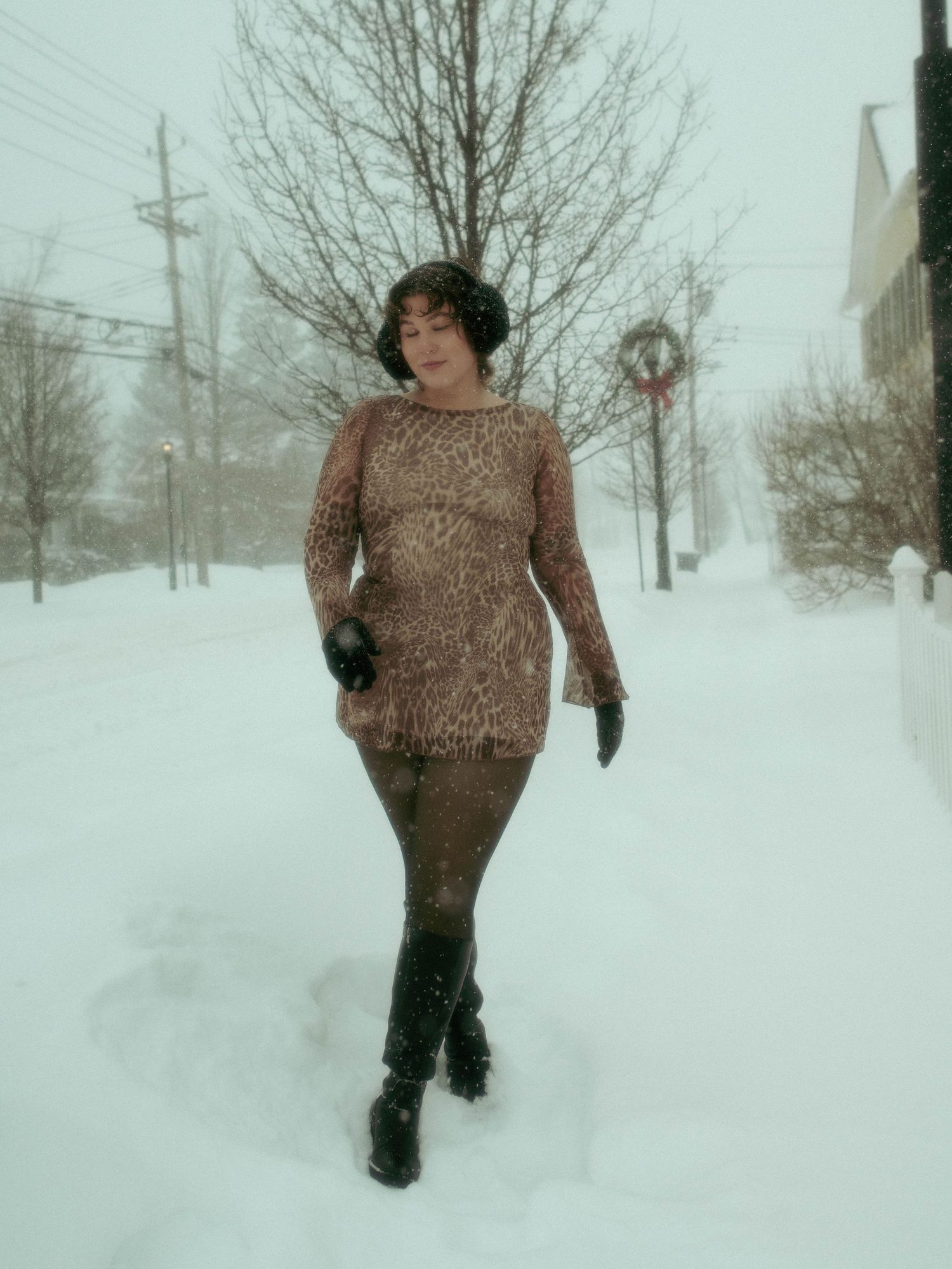Person wearing brown sweater walks through deep snow during winter storm with utility poles visible in background.