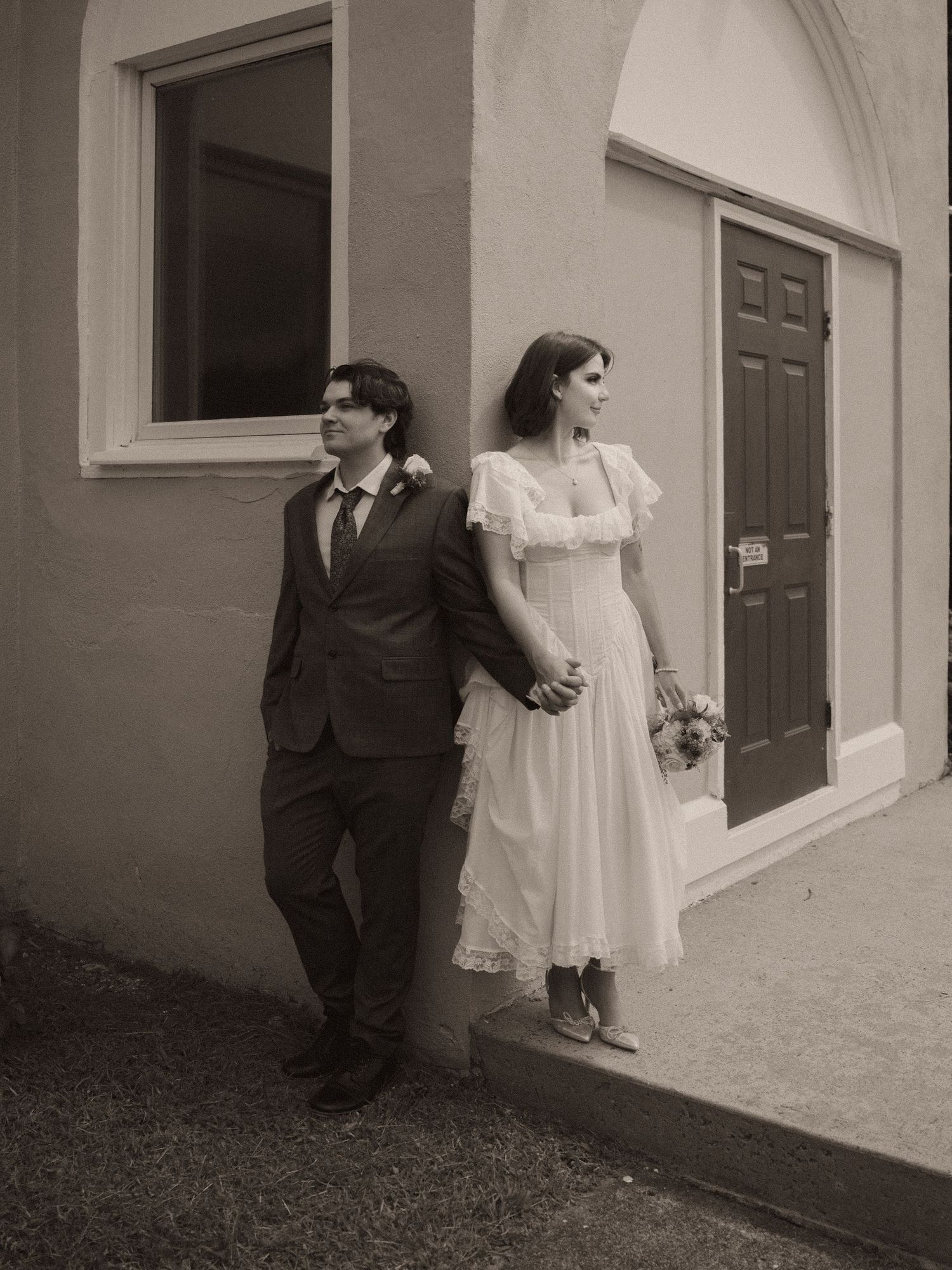 Bride and groom hold hands leaning against building wall in classic black and white wedding photo.
