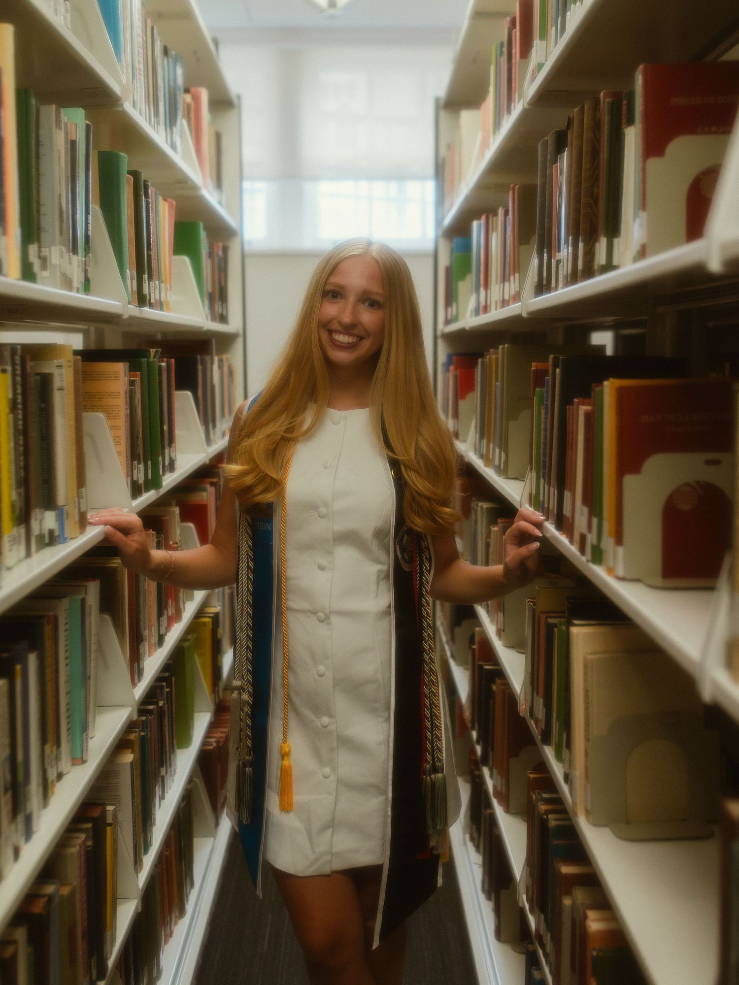 Smiling blonde woman in white dress holding graduation regalia between library bookshelves.