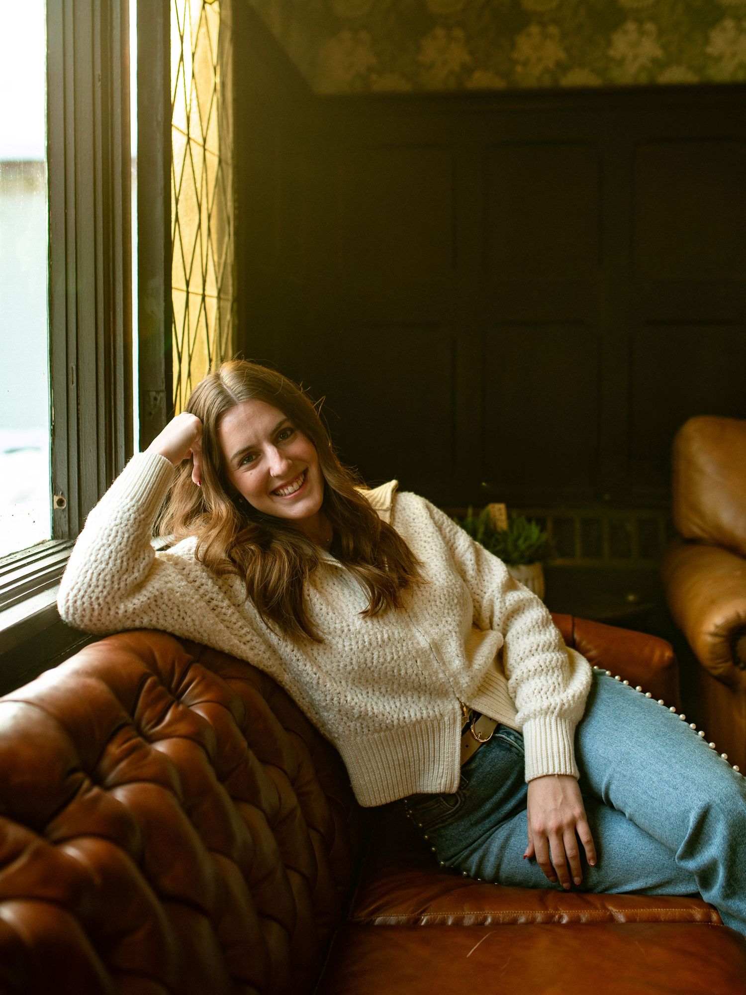 Person relaxing comfortably on a brown leather couch in a sunlit room wearing a cozy cream sweater.