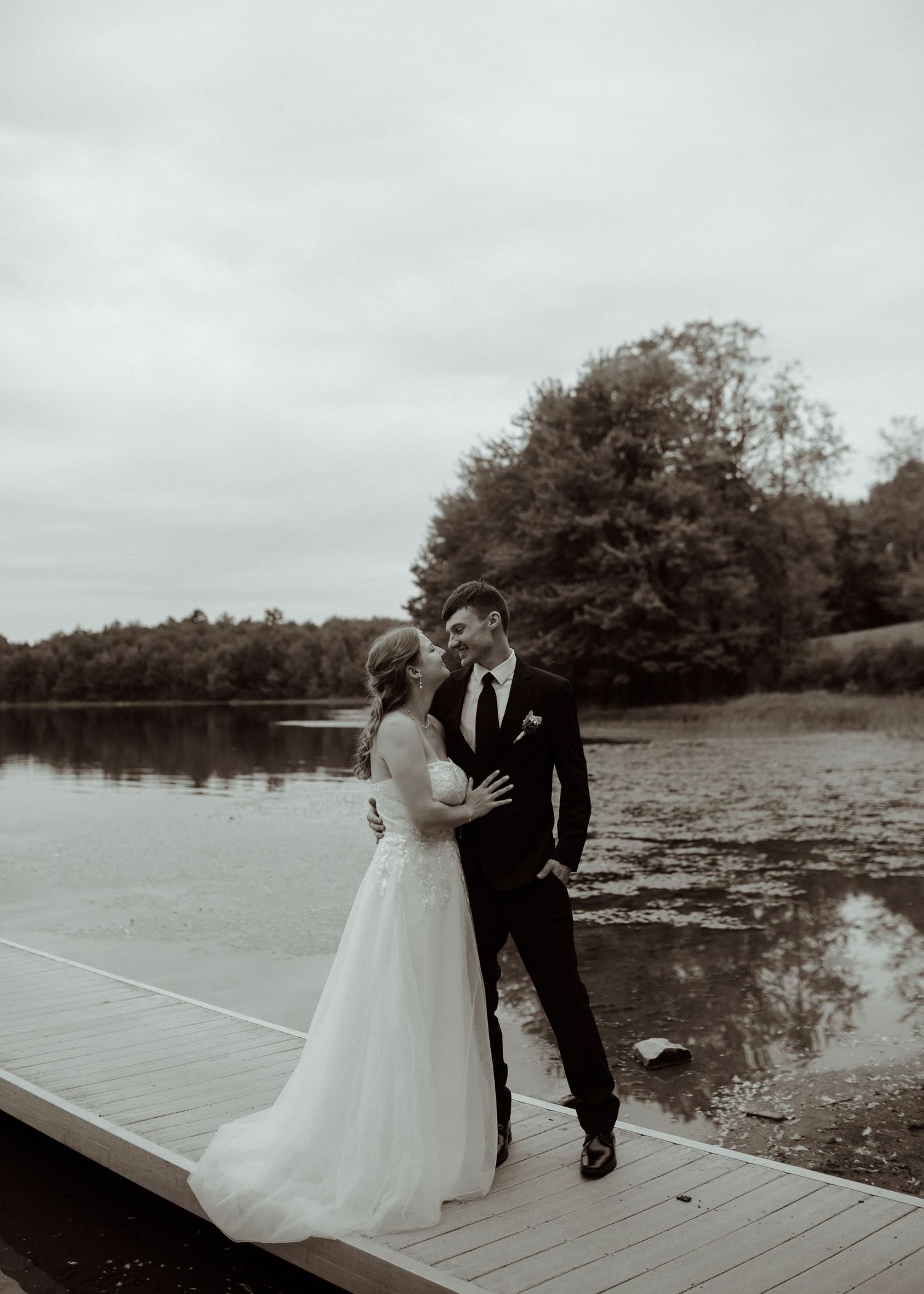 Couple in wedding attire poses together on a wooden dock by a lake.