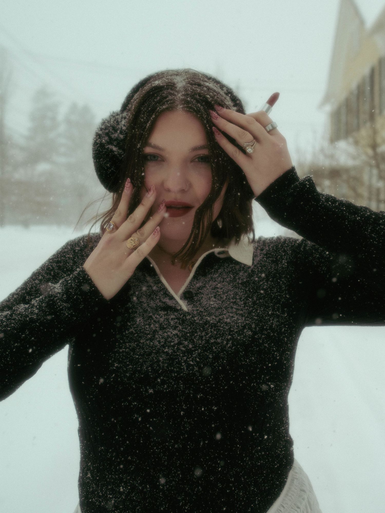 A person in a black winter coat playfully covers their face while standing in falling snow.
