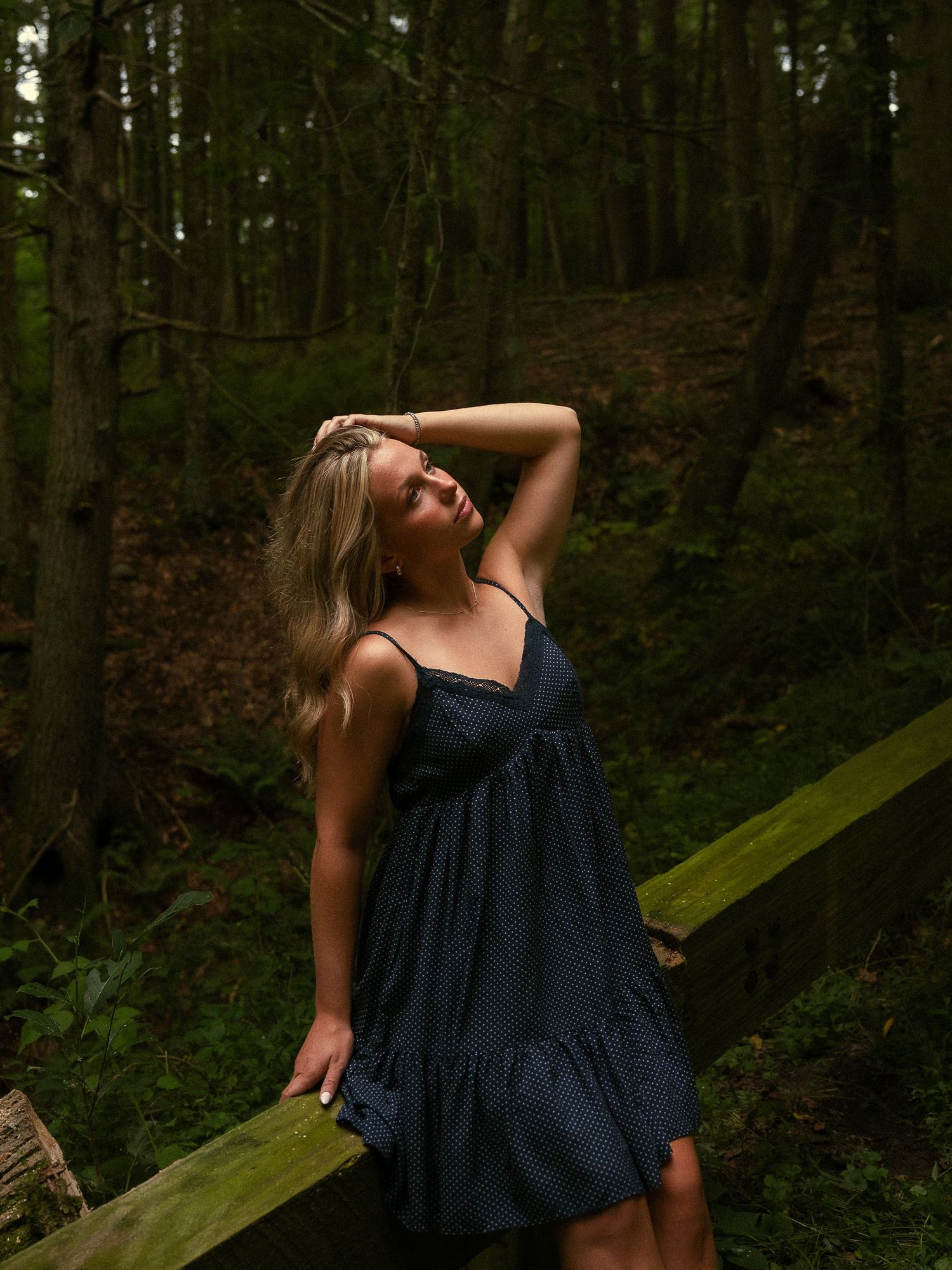 A blonde woman in a navy blue mini dress poses gracefully in a lush green forest setting.