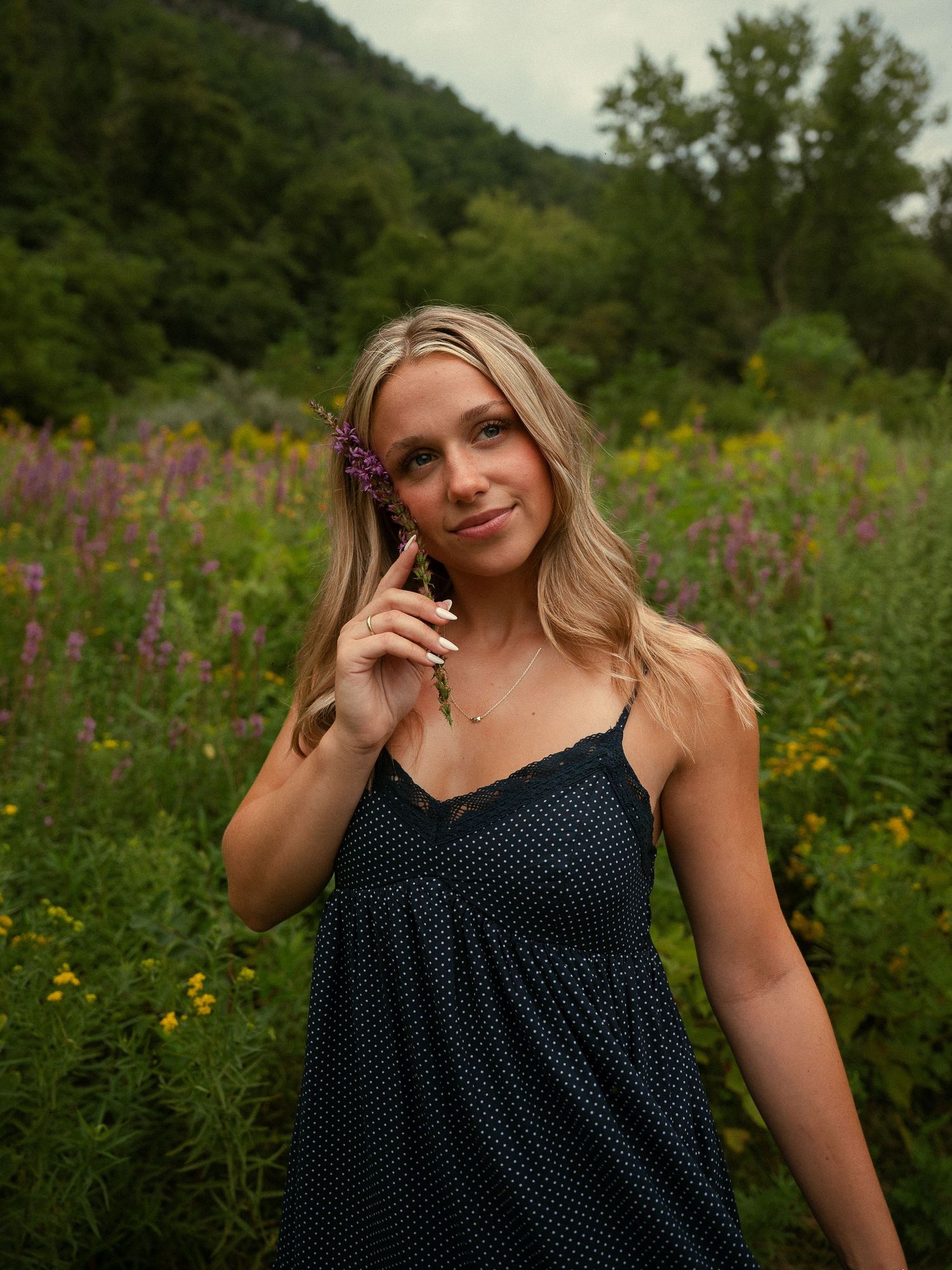 A young blonde woman in a navy polka dot dress holds purple wildflowers in a colorful meadow with green hills behind her.