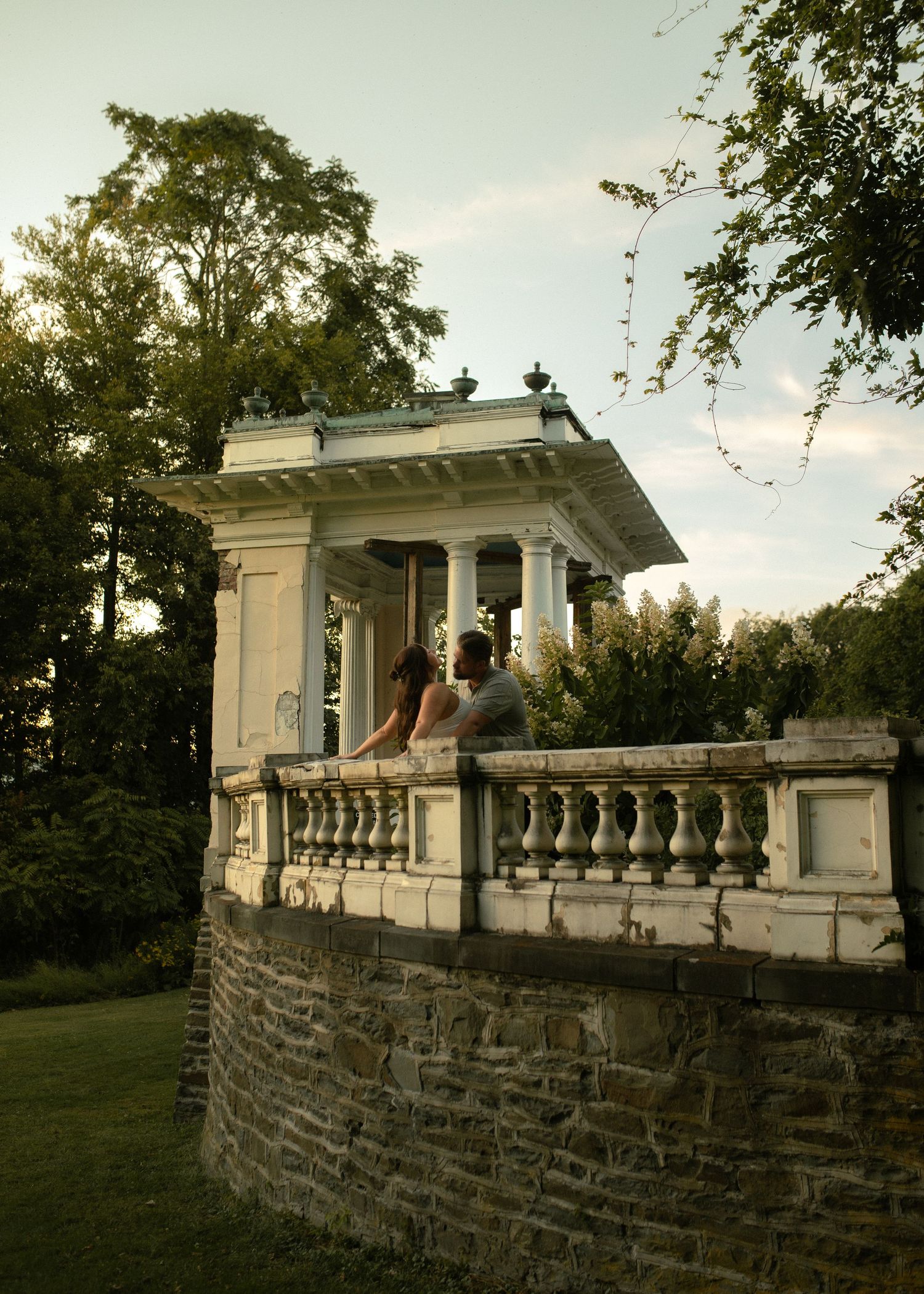 Classical white stone gazebo with columns photographed at sunset in a garden setting.