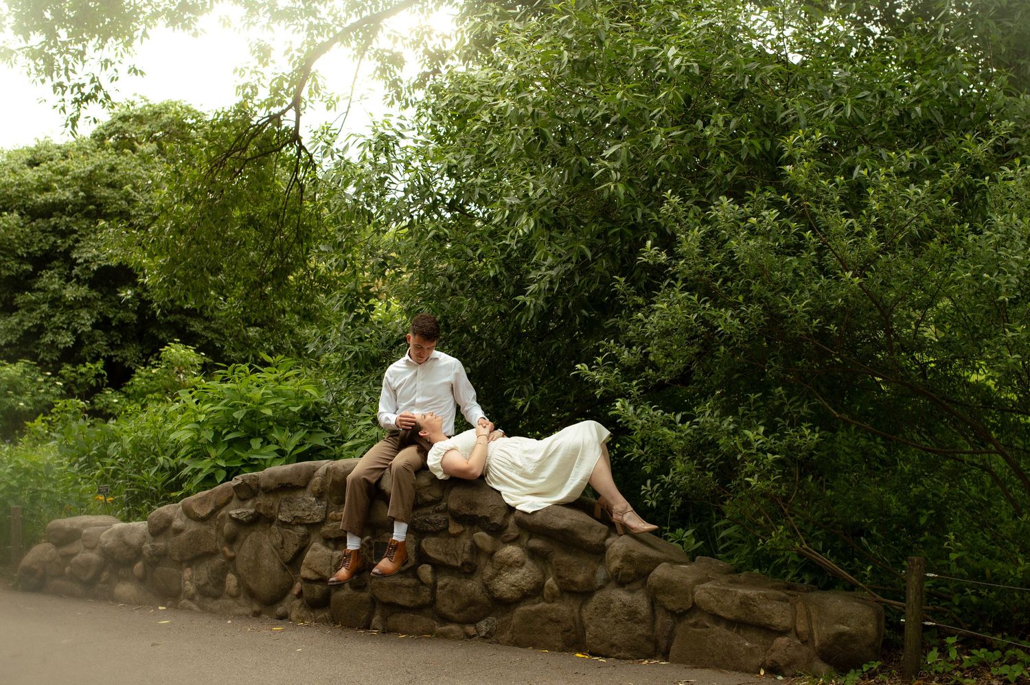 A couple in white clothing poses romantically on a stone wall surrounded by green foliage.