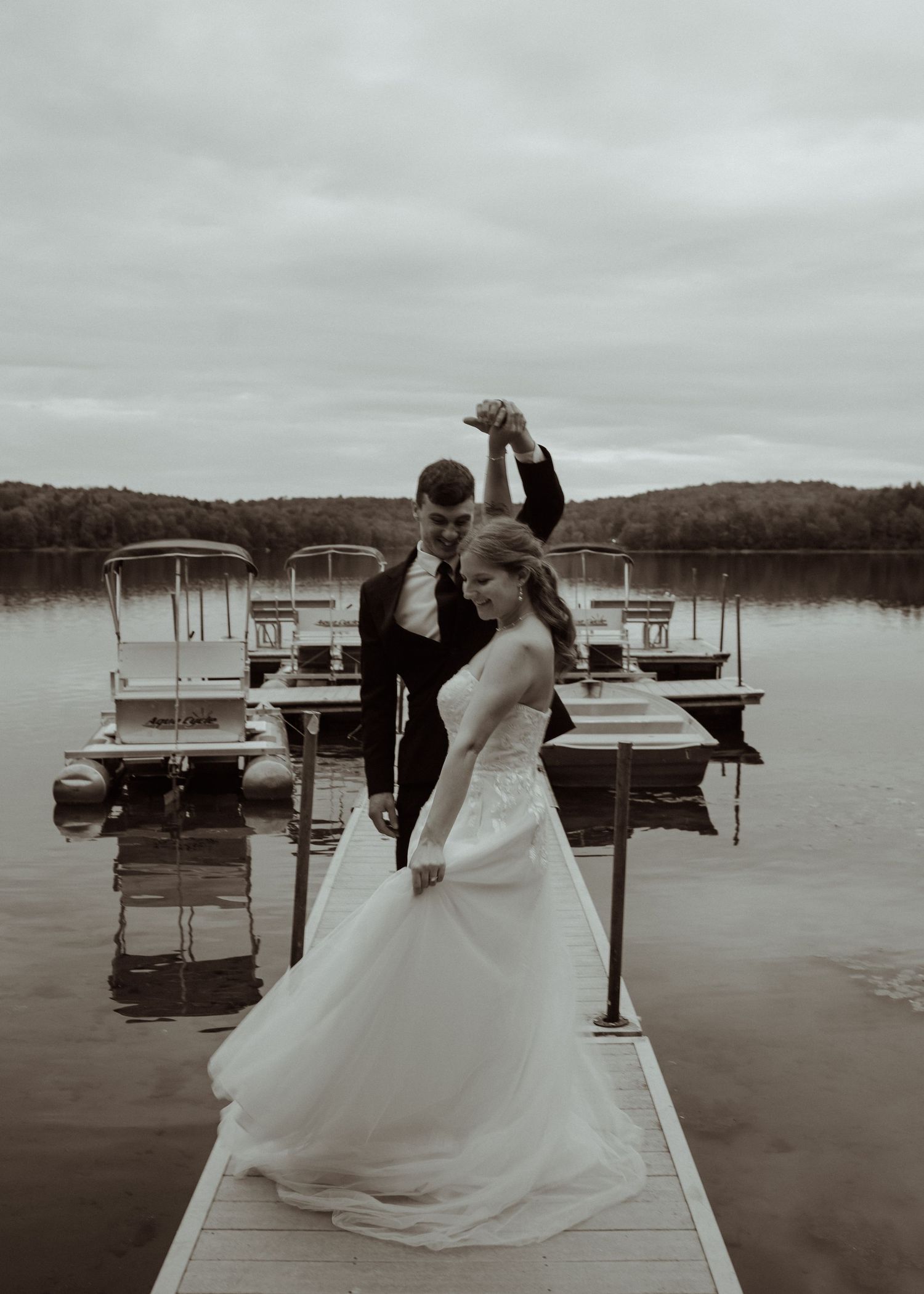 Couple in wedding attire dances on a dock with boats in the background.